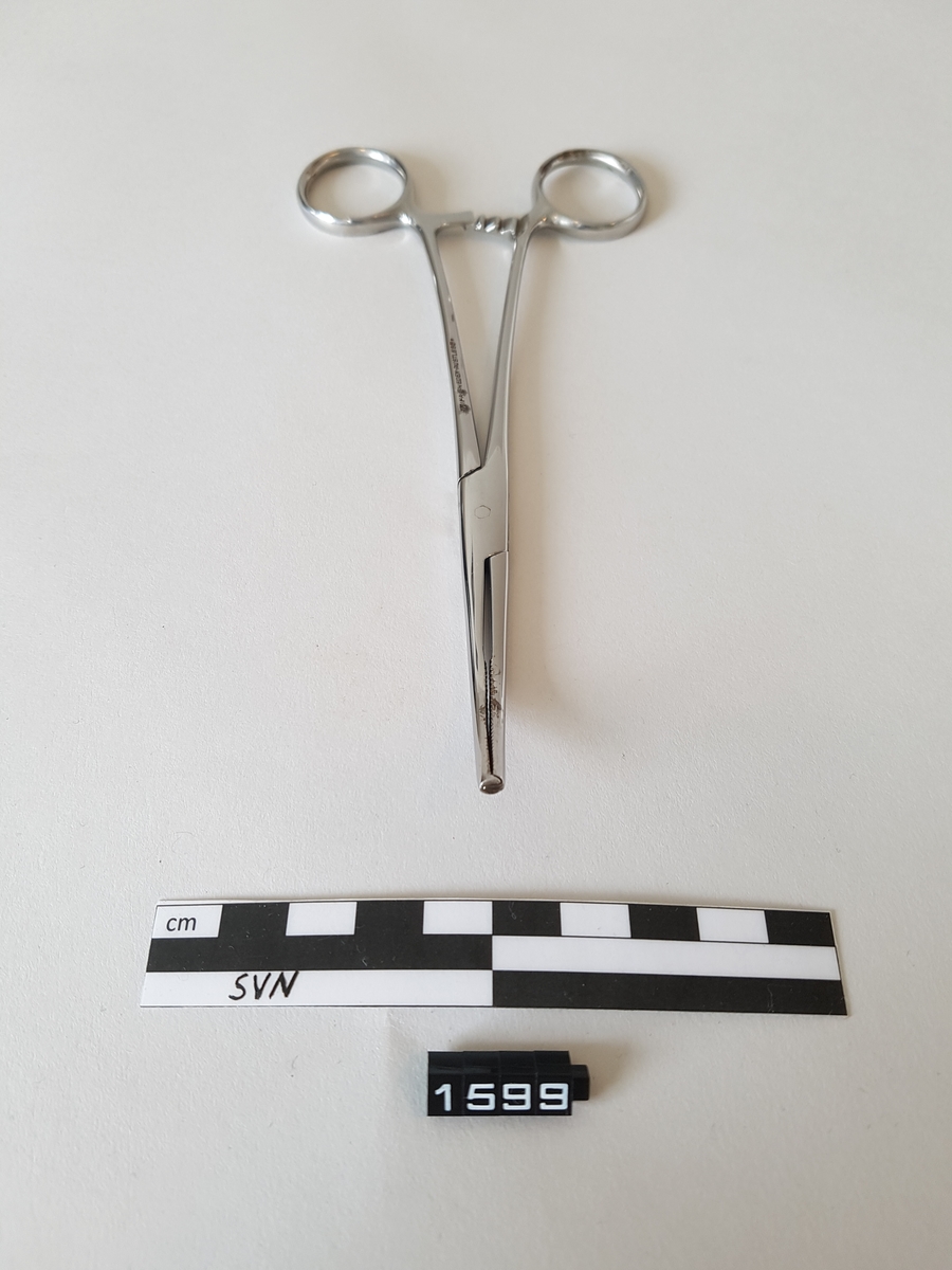 Gjenstanden er en kirurgisk bøyd klemme. 
Klemmen er utformet som en saks, men i stede for skjærekniver er det to flater som møtes og klemmes sammen.
Flatene som møtes har serrateringer, riller. ytterst på klemmen har flatene tenner som går i hverandre.
To tenner på en side og en tann på den andre siden.
Gjenstanden har fingerhull for manipulering av objektet.
like over fingerhullene er det en låsemekanisme med en hake på den ene delen og flere motstående haker på den andre delen
Instrumentet kan låses ved hjelp av disse og hvor mye press det legges på tangspissen bestemmes av hvilken av mothakene 
det låses mot.