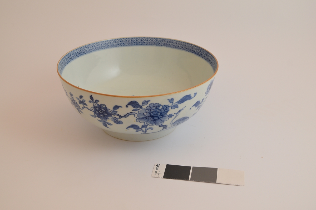 En hvit porselensbolle som er dekorert med blomster på utsiden av bollen og en blå mønstret kant langs toppen på innsiden av bollen som er ca. 2 cm høy. Det er i tillegg in gul stripe langs toppen av veggen til bollen.