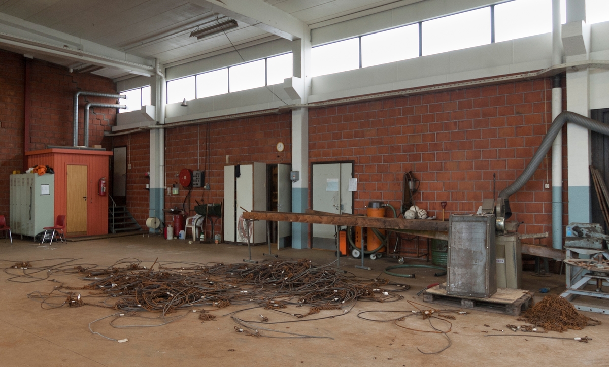 Fra Skiensvassdragets fellesfløtningsforenings verksted i Skien.  Fotografiet viser en hall med betonggolv og bærekonstruksjoner av prefabrikerte betongelementer, og med vegger som er kledd med teglfliser og har lysinnsig fra ei vindusrekke oppunder gesimsen.  Diverse skap og arbeidsbenker står langs veggene.  På golvet ligger det en del rusten vaier, som antakelig skulle repareres med sikte på gjenbruk. 