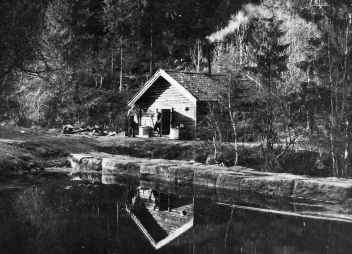 Den såkalte «Kringlekoia» i Løvenskiold-Vækerøs skog i Nordmarka.  Koia ligger ved en fløtingsdam, og vi skimter damkrona på steindammen på skrå i forgrunnen.  Gavlen på den laftete koia speiles i vannflata.  Den har inngangsdør sentralt på gavlen.  Da bildet ble tatt stod den åpen.  På hver side av den stod det to trekasser, antakelig for proviant.  To unge menn befant seg foran døråpningen, den ene med bar overkropp, antakelig fordi han var i ferd med å vaske og barbere seg etter ei natts søvn.  Koietaket var tekket med krumtegl.  Fra et langt jernrør gjennom mønepartiet stiger det opp røyk, noe som tyder på at det var ild i koieovnen.  Fotografiet ble tatt på forsommeren 1942. 