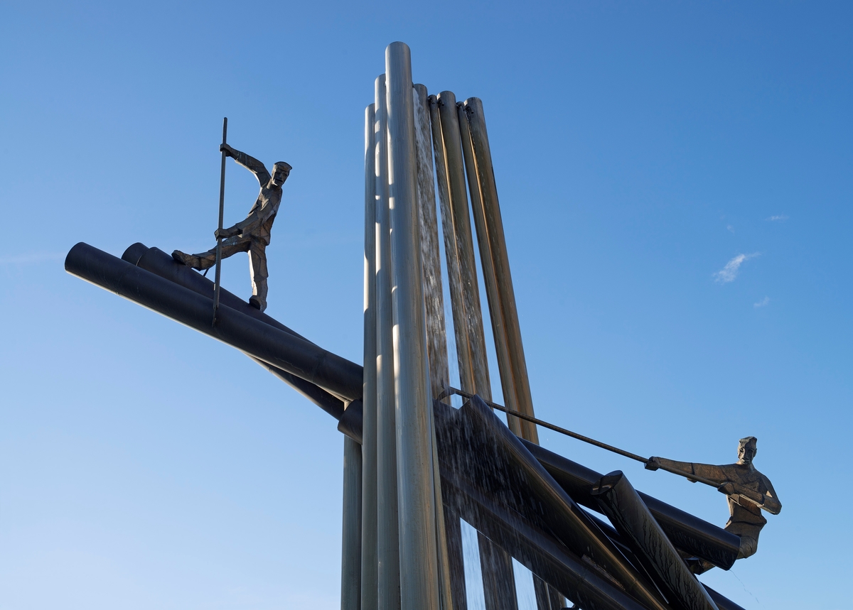 Detalj fra fløtermonumentet i Skien.  Det består av ei gruppe søyler som bærer en montasje som antakelig en tømmerhaug i et fossefall, der to fløterskikkelser forsøker å løsne stokkene ved hjelp av fløterhaker.  Dette utsnittet viser den øvre delen av skulpturen med blå himmel som bakgrunn.  Vi ser to fløtere i arbeid.  Den ene skyver en tømmerstokk han har slått hakespissen i, den andre drar.  Idéen om å sette opp et fløtingsmonument i Skien ble lansert tidlig i 1990-åra.  I 1991 mottok Skiensvassdragets Fellesfløtningsforening en søknad om penger til prosjektet fra Skien kommune, som opprinnelig ville plassere monumentet ved den den gang nye gang- og sykkelstien over fløtingsorganisasjonens tomt.  Samme år vedtok Skiensvassdragets Fellesfløtningsforening å bidra med 50 000 kroner til prosjektet.  De utfordret også Lågen-Telemark tømmermåling.  Målet var at monumentet skulle stå ferdig til 100-årsjubileet fra Bandak-Norsjø-kanalen i 1992.  Monumentet ble reist på nordre del av Smieøya. 