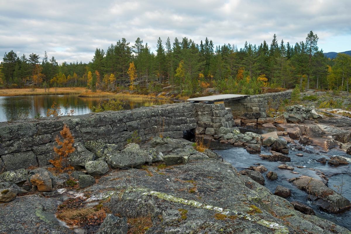 Dammen ved utløpet av Øvre Bjørvatn eller Øvre Bjorvatn [navneform brukt i dokumenter fram til midten av 1900-tallet] i Tinn i Øst-Telemark.  Bildet viser hvordan dammen er plassert på et sted der vann fra den nevnte lille sjøen renner ut over ei bergflate.  Dammen er murt i tuktet naturstein.  Den har ett løp, som sto åpent da dette fotografiet ble tatt, men som antakelig ble stengte med «nåler»,  plank som ble stukket tettstilt ned på oversida av dambrua, slik at de ble presset mot en terskelbjelke på botnen og brukanten.  På den ene sida av damåpningen, og vel så lavt, er det lagd en liten, kvadratisk åpning.  Denne må antakelig ha fungert som «botnluke», altså som uttappingskanal når det (i lavvannsperioder) skulle utføres reparasjonsarbeid på damkonstuksjonene.  Fra gammelt av var det en steinfylt tømmerkistedam ved Øvre Bjørvatn.  Fotografier fra slutten av 1930-åra viser at tømmerkistene da var svært dårlige (jfr. SJF-F. 005450, SJF-F. 005452 og SJF-F. 005453).  Den fine natursteinsdammen på dette fotografiet ble bygd i 1946.  Smeden og gråsteinsmureren [Karl] Marius Kristiansen ble tilbudt 5 000 kroner for å mure en ny dam.  Han hadde tidligere gjort seg positivt bemerket, blant annet i arbeidet med Rauatunnelen lengre nede i vassdraget i slutten av 1920-åra.  En kultursti ved Rauatunnelen forteller følgende om Kristiansen og hans arbeidsinnsats:  «Karl Marius Kristiansen (1886-1972) var frå Våle i Østfold. Han kom til Hovin i byrjinga av 1920-åra, etter å ha vore på mange anlegg, t. d. hadde han arbeidd i Sulitjelma gruver. Marius var flink til all slags arbeid, særleg flink var han som smed og murar. Han blir omtala som arbeidsnarkoman, og sjølv om det var lange arbeidsdager og tungt arbeid, kunne han ikkje forstå folk som la seg ned til å sove når dei hadde ei fristund, det var så mykje anna å bruke tida til. Det er sagt om han at han aldri forhandla om betre løn, han sette heller opp tempoet enno meir dersom akkorden var låg. Alt medan Marius lede vart han omtala som ein legende, t. d. ville han ikkje ta i mot alderstrygd, etter hans meining skulle det arbeidast for pengane. »

Fløtinga i Rauavassdraget begynte gjerne i midten av mai og pågikk tre-fire uker.  Deretter ble tømmeret fra vassdragets nedslagsfelt buksert mot Tinnoset med slepebåten "Maar".  Utover i 1960-åra krympet fløtinga i sidevassdragene noe, for det ble stadig vanskeligere å rekruttere folk til sesongarbeid langs vassdragene samtidig som mulighetene for lastebiltransport av tømmer ble stadig bedre.  I Rauavassdraget oppsto det i tillegg spesielle problemer knyttet til slitasje på betongforblendinga i Rauatunnelen, nederst i vassdraget.  1968 var det siste året det var fløting her. 