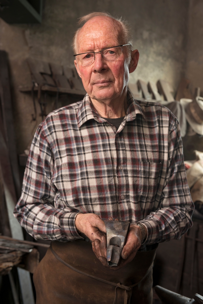 Smed Oddbjørn Knudsen (f. 1935), fotografert i smia med ei ferdig, men uskjeftet merkeøks i hendene.  Bildet er tatt i Knudsen-smia på Veiavangen i Mjøndalen (Nedre Eiker), der Oddbjørn er tredje generasjon øksesmed.  Han er kledd i rutete flanellskjorte og et lærforkle som dekte mage, hofter og lår under arbeidet i smia, hvor det tidvis kunne fyke en del gnister fra essa og fra glødende metall som skulle formes. 