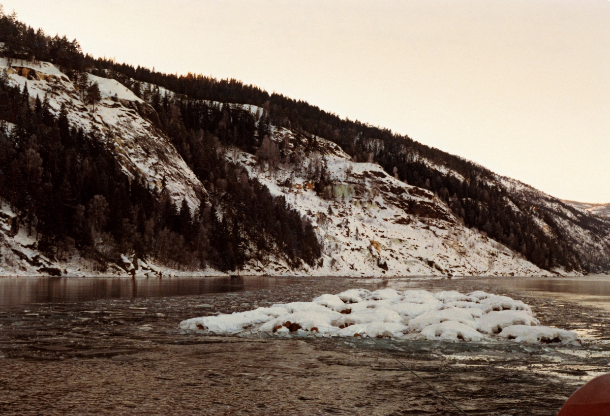 For mannskapene som slepte tømmer på innsjøene i Skiensvassdraget var det om å gjøre å få mest mulig virke ned til Skien før vinteren kom med kulde og is.  Dette fotografiet er tatt fra akterenden av slepebåten Axel, som trakk tømmer over Bandak, Kviteseidvatn og Flåvatn, idet båten passerte Venheimskottet i ytterenden av Flåvatn.  Fotografiet viser slepet, buntet tømmer som var knyttet sammen med jernvaiere.  På denne kalde høstdagen var buntene snødekte, men vassdraget var fortsatt åpent, slik at virket kunne trekkes ned til opptaksplassen ved Kåsa (Saltevje) i Nome. 