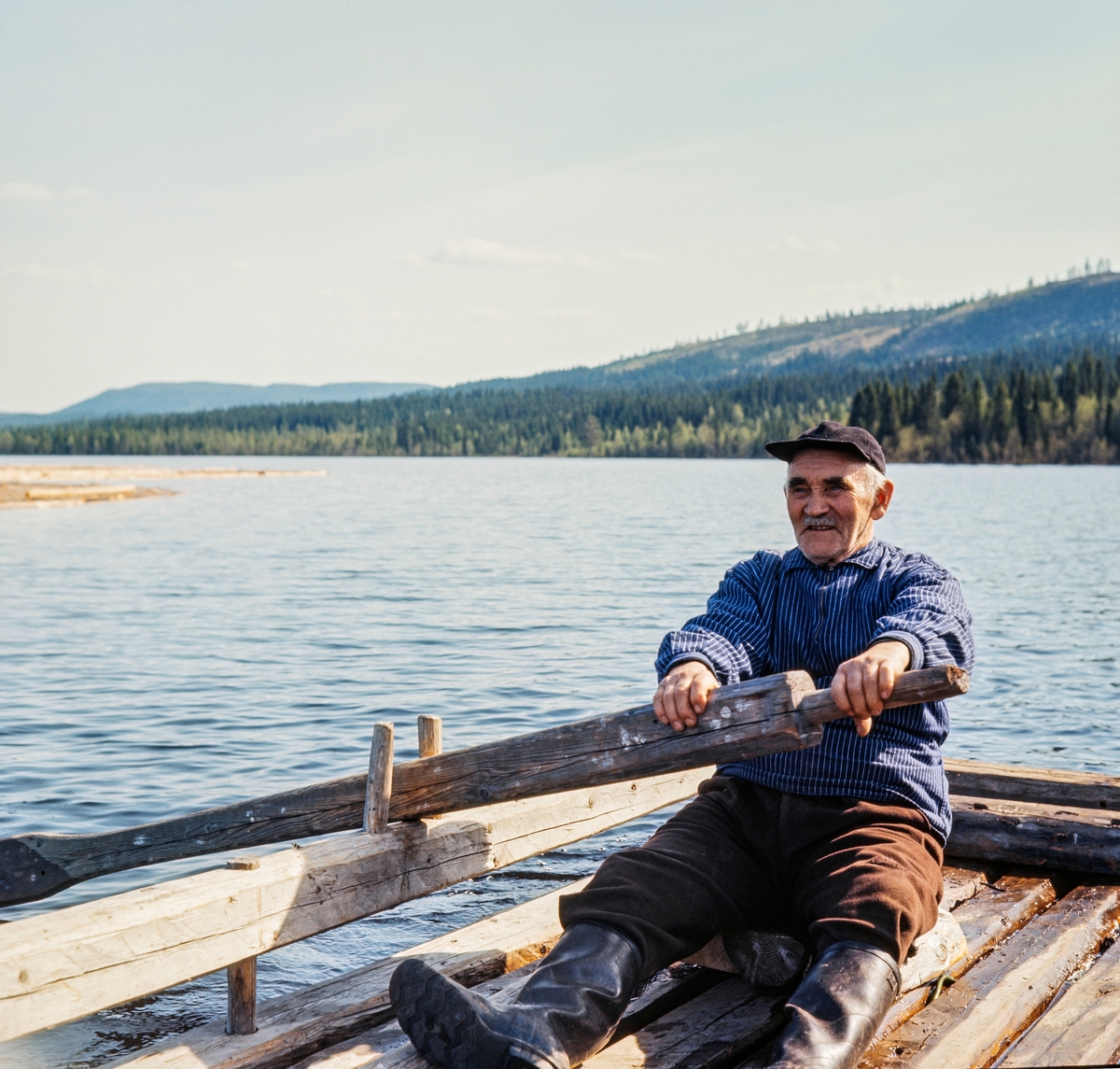 Johan Sjølie ror spilleflåte i en av de to Ulvsjøene i Trysil.  Sjølie sitter på ei steinhelle i et av hjørnene på flåten med et godt grep om ei kraftig åre, som ligger an mot tollepinner på en av sidestokkene på flåtekonstruksjonen.  Fløteren hadde brune vadmelsbukser, lyseblå busserull med kvite striper og svart skjoldlue på hodet.  På beina hadde han svarte gummistøvler.  I bakgrunnen ser vi vannspeilet (med en tømmerbom til venstre) og omkringliggende delvis skogdekte åser.  En av de tre andre karene som arbeidet sammen med Sjølie på denne flåten satt antakelig i andre hjørner og rodde på samme måte når flåten skulle flyttes uten etterhengt tømmerbom.  Da dette fotografiet ble tatt rodde fløterne sannsynligvis for å hente en ny tømmerbom. 

Johan Sjølie var fløtersjef for Ena og Ulvsjøene fra 1938 til 1965.  Han skal ha vært en rolig, sindig og pålitelig kar, som var godt likt av så vel fløtere som skogeiere.

Litt generell informasjon om spillflåtefløtinga på Ulvsjøene finnes under fanen «Andre opplysninger». Ulvsjøen. Hedmark.