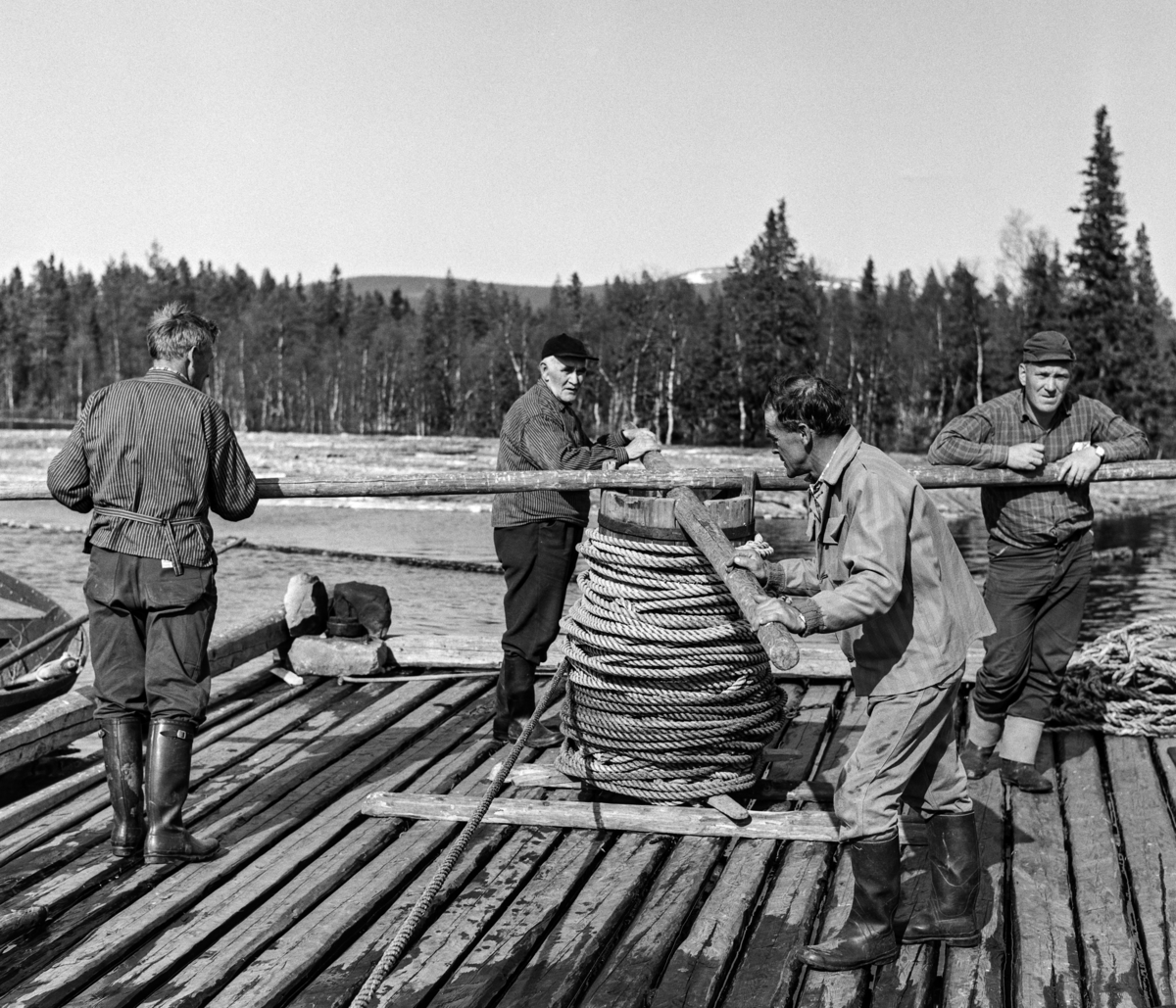 Arbeidslag på spillflåte på en av Ulvsjøene i Trysil i mai 1969.  Fotografiet er tatt mot sentrum av den noenlunde kvadratiske spillflåten, der karene beveget seg på.  I sentrum av denne er det reist en cirka en meter høy sylinder, som ble kalt «tønna».  På toppen av denne roterbare konstruksjonen er det lagt to trestenger - «årer» - i kryss.  Dette er et gangspill med ei cirka 200 meter lang trosse med et anker - kalt «krabba» - i ytterenden.  Krabba ble rodd framover i sjøen, så langt trossa rakk, og sluppet ned på botnen, hvor den forhåpentligvis festet seg.  Deretter kunne karene «spille» - dreie gangspillet rundt og rundt - slik at flåten og en bakenforliggende tømmerbom sakte men sikkert ble trukket mot ankringspunktet.  Når de nådde fram dit, ble krabba hevet og rodd videreframover i den retningen man ønsket å ha tømmereet. En av robåtene som ble brukt ved slike forflytninger skimtes til venstre i bildet.  På bildet ser vi Halvor Sjølie (til venstre med ryggen mot fotografen), Johan Sjøli (med skjoldlue), Egil Torfinn Bakken (i forgrunnen) og Magne Neby.

Litt generell informasjon om spillflåtefløtinga på Ulvsjøene finnes under fanen «Andre opplysninger». Ulvsjøen. Hedmark.