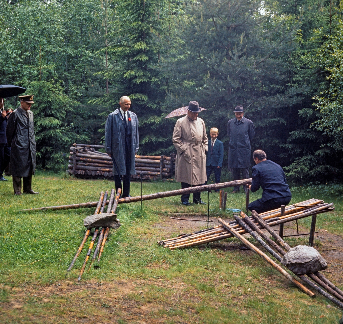 Omvisning i friluftsmuseet på Prestøya i forbindelse med at kong Olav V (1903-1991) åpnet Norsk Skogbruksmuseums nybygg 22. juni 2014.  Omvisningen ble holdt etter at åpningsseremonien og en lunsj var avviklet inne i museet.  Konservator Tore Fossum (1926-2017) hadde mobilisert herredsskogmestere fra Hedmark og fiskere fra Sølensjøen i Rendalen til å bemanne koier og anlegg.  Her demonstrerer museumsbestyreren fallstokk.  Tilskueren lengst til venstre (i regnfrakk og uniformslue) er fra kongens følge.  Deretter følger sekretæren i museets styre, Wilhelm Matheson (1910-2001), kongen, Torstein Fossum (konservatorens sønn) og en mann vi foreløpig ikke kjenner identiteten til.