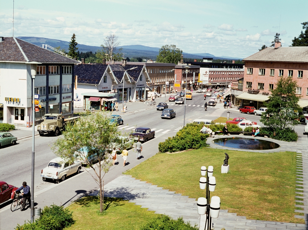 Storgata i Elverum sentrum (Leiret) sett fra et vindu i 2. etasje i kommunens rådhus sommeren 1969. I forgrunnen ser vi den lille grønne parken på forsida av bygningen, der det også var et lite, rundt basseng foran inngangen til biblioteket. Ellers ser vi krysset mellom Storgata og St. Olavs gate. Bebyggelsen langs gateløpene var preget av stilidealer fra gjenoppbyggingsperioden etter 2. verdenskrig. Bare ett av de gamle trehusene mot tettstedets hovedgate – villa «Bæk» fra 1875 – sto fortsatt, men var ombygd til forretningsformål.