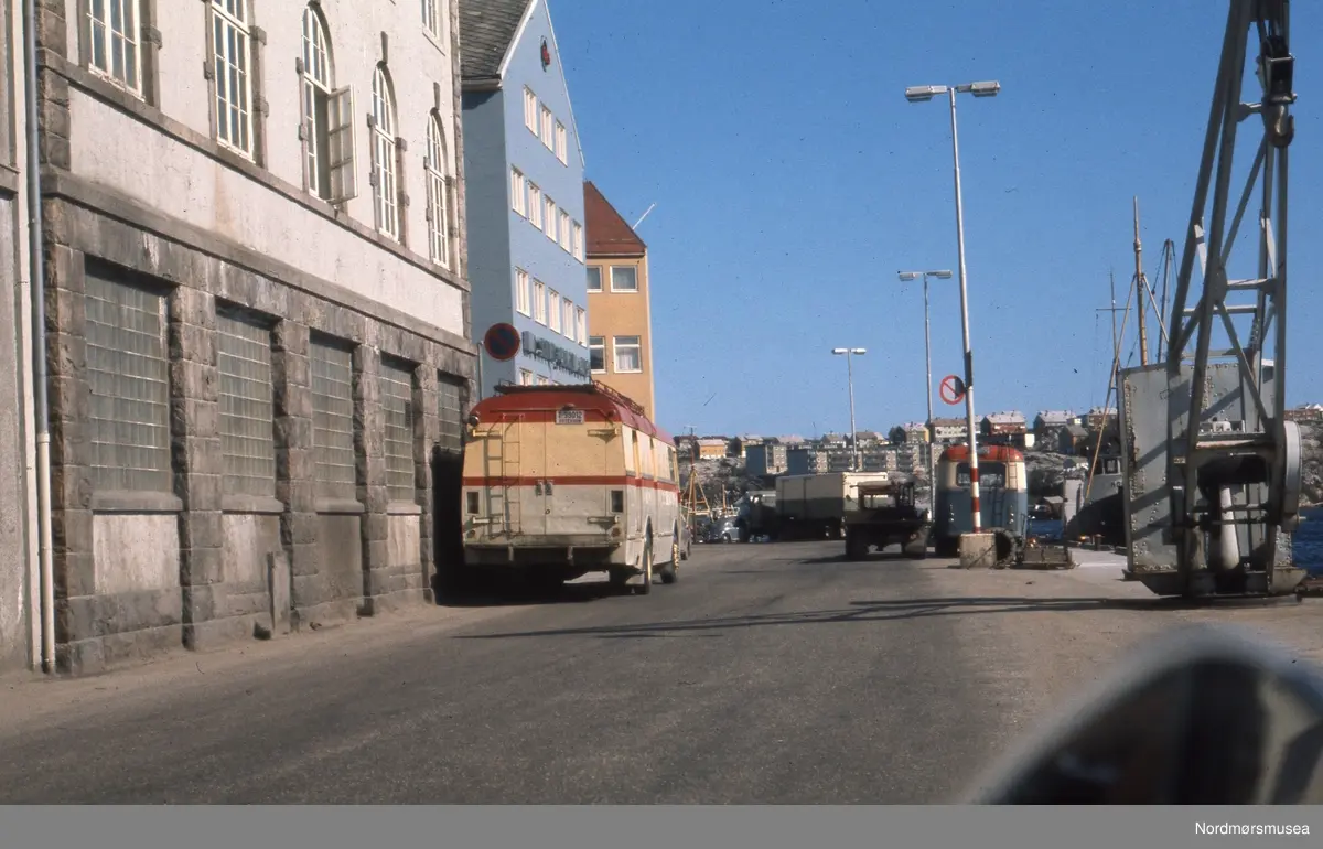 Merk "Meieriveggen" fra 1917 som ble bevart da Amfi Storkaia brygge bygde om husrekka på Storkaia/Arnulf Øverlands gate. Nå 2018 er Coop Mega innenfor her.  ---  Meieriene var tidligere en viktig losse- og lasteplass for lokale rutebiler. Melk ble levert i spann fra bøndene, og konsummelk ble fraktet fra meieriet til alle kolonialbutikkene i distriktet. Midt på bildet ser vi bilde bakfra av en kombinertbuss fra Smøla Billag. Registreringsnummeret T-99012 er lesbart. Dette var en Ford 1964-modell, og den har selskapets karakteristiske farger, lysgrå nederst, gul overdel med røde striper og tak. Til høyre på kaikanten ser vi en buss fra Kristiansund Frei Billag, i blågrå, rødt og gult. I bakgrunnen ses en semitrailer i grønt med lys overdel. Denne tilhørte fra Averøy Billag. (Info: Sveinung Berild). --  Foto fra Meieriet på Kirkelandet i Kristiansund, her sett fra Storkaia. Fotografier og karttegninger, fotografert eller samlet sammen av Arkitekt MNAL Kristian Sylthe (f. 18.06.1926) ved Kristiansund kommune i sitt virke som kommunearkitekt (senere næringssjef) samt som sekretær i Oljeutvalget fra begynnelsen tidlig 1970. Samlingen kan i hovedsak dateres mellom 1945 til 1980. Samlingen er gitt i gave av Kristiansund kommune. Fra Nordmøre museums fotosamlinger.