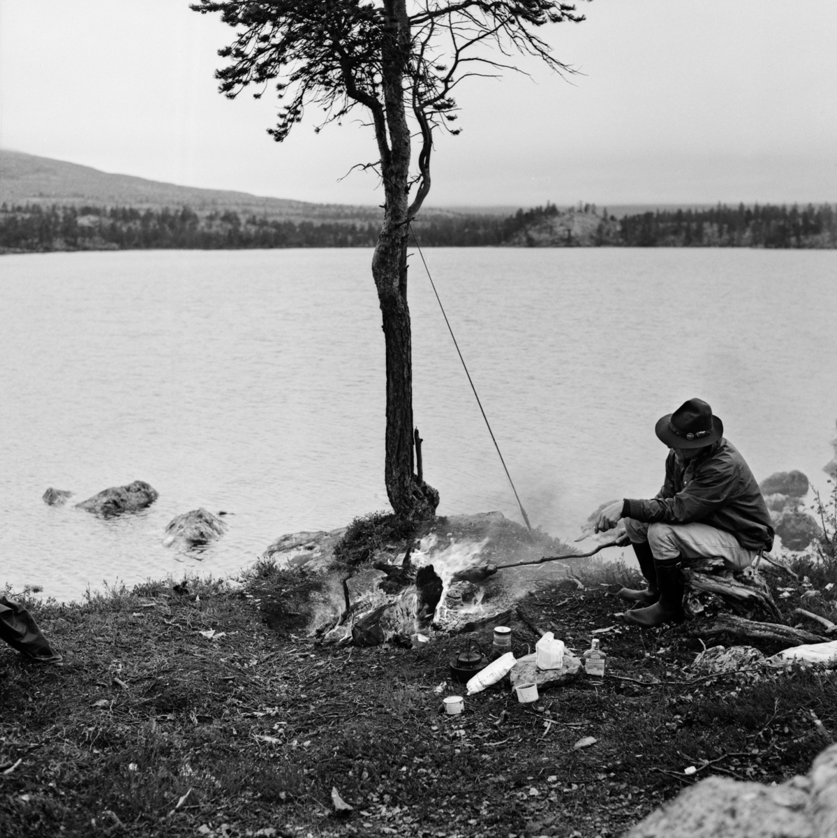 Grilling av fisk vet et av vatna i Femundsmarka.  Fotografen er tatt på stranda, der fiskeren hadde tent seg et bål hvor han grillet en av fiskene han hadde fått over åpen ild.  Han satt på en stubbe med fisken tredd inn på ei grein.  På bakken foran mannen og bålet ser vi en del andre nærings- og nytelsesmidler fiskeren hadde med seg, blant annet en kaffekjel og ei brennevinsflaske.  Mannen hadde ei lys bomullsbukse, mørk vindjakke og en vidbremmet hatt på seg.  På beina hadde han langskaftete gummistøvler.  Bak bålet hadde han reist fluestanga si mot ei lita furu som vokste like ved vannkanten.  Fotografiet ble tatt i 1966. 