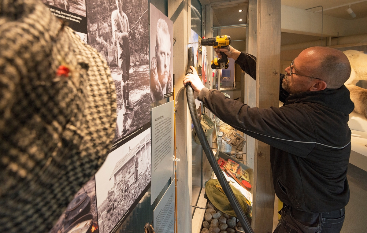 Eskild Midtlie borer hull til et oppheng i Norsk Skogmuseums nye basisutstilling «Tråkk i mangfoldig natur» våren 2014.  Han arbeider med drill i høyre hånd og holder en støvsugerslange i den venstre, for å unngå at flis fra boringa faller ned i den underliggende montasjen.  Fotografiet er tatt ved en montasje som handler om fritidsfiske.  Her er det montert fotografier av sportsfiskere i ulike situasjoner, og et portrett av polarhelten Fridtjof Nansen, som var et forbilde for mange også når det gjaldt friluftsliv og fiske.  Montasjen inneholder ellers et par fiskestenger, fiskeskry i ulike materialer og klesplagg som det har vært vanlig å bære under fisket.  Montasjen har følgende hovedtekst: «Fritidsfiske.  Gleden over å få fisk, avkoblingen og naturopplevelsen er de viktigste drivkreftene i fritidsfiske.  På midten av 1800-tallet oppdaget den engelske overklassen Norge som et sted for rekreasjon, fritidsfiske og fjellvandring.  Fridtjof Nansen ble gjennom sin bruk av naturen et forbilde for mange friluftsfolk og fritidsfiskere.  Fritidsfiske er i dag en av de mest utbredte friluftsaktivitetene i Norge.  Nesten halvparten av voksne nordmenn fisker i løpet av året, og barn lærer gjerne å fiske av de voksne.  Vi fisker mest ørret og laks, men andre arter øker stadig i popularitet.  I dag slippes mye av fisken ut igjen, såkalt «fang og slipp», som er en omdiskutert praksis. »