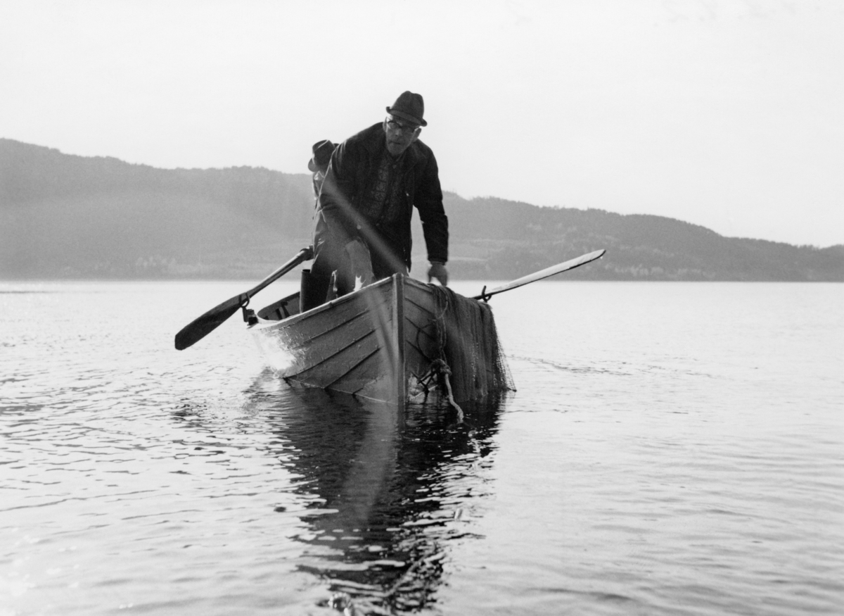 Forberedelser til notkast etter krøkle ved Langodden i Furnesfjorden i Mjøsa.  Fotografiet viser en robåt, som krøklenota nok skulle kastes fra.  Bildet ble tatt idet båten seg ut fra land med en mann stående i baugen, der nota lå klar på båtripa, og en mann som var i ferd med å få årene på plass i kipene bakenfor.  I vannet innenfor båten aner vi et nottau, som det antakelig var en mann på land som holdt i, og som etter hvert skulle brukes ved inndraing av redskap og potensiell fangst. 

Notkastet karene på dette fotografiet var i innledningsfasen til, var nok et rekonstruksjonsforsøk knyttet til noe som hadde vært et tradisjonelt vårfiske i Furnesfjorden, antakelig fram til disse karenes barndom.  En del større garder hadde hevd på å drive såkalt «langrevfiske», en ferskvannsvariant av linefiske, utenfor sine strandlinjer.  I dette fisket brukte man levende krøkle som agn på langrevens kroker.  Krøkla ble fanget i nøter, som den vi ser på dette fotografiet, når den kom inn mot strendene for å gyte om våren. Denne formen for fiske opphørte etter at det ble forbudt å bruke levende agnfisk.  Karene på fotografiet hadde følgelig neppe vært med på notdraing etter krøkle.  De hadde imidlertid med seg en 90-årig veteran, som nok kunne fortelle hvordan det ble gjort (jfr. SJF-F. 007189).  Fiskeredskapen var antakelig utlånt fra boet etter brødrene Hans (1900-1966) og Martin (1904-1960) Storihle på garden Stor-Ile i Furnes.  Fra denne garden ble det drevet et omfattende fiske i Mjøsa, til dels med sikte på videresalg av fisk til kjøpmenn på Hamar og i Oslo.  Garden har fortsatt fiskerhytte på Langodden. 