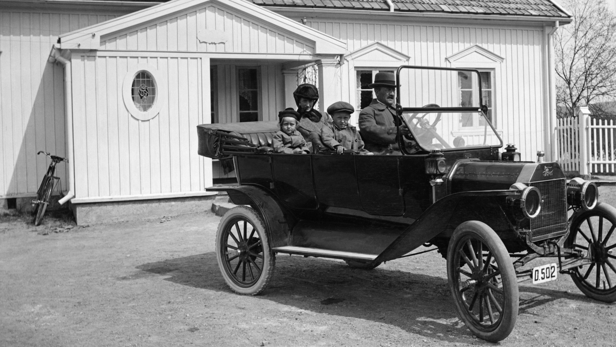 Familien Astrup fotografert i en T-Ford foran våningshuset på garden Haug i Våler i Hedmark.  T-Forden var en lett, forholdsvis enkel bilkonstruksjon, med et platekledd karosseri som bestod av et rammeverk med trespanter. Det ble kledd med et stoff kalt pegamoid.  I oppholdsvær sommerstid, som her, kunne kalesjeoverdekningen foldes tilbake.  Ved rattet i den åpne bilen satt familiefaren Carl Astrup iført hatt og frakk.  Ved sida av ham ser vi sønnen Christian.  I baksetet satt dattera Anna Helene og kona Ludovica, som var født på Kjellmyra i Åsnes som datter av gardbruker og landhandler Georg Kristian Fleischer og kona Anna.  Carl og Ludovica Astrup kjøpte garden Haug med underbrukene Klyperud og Berg, samt seks skogteiger i Gåsvassdalen, i 1906.  Deretter satte de i stand bygningene på eiendommen.   Når det gjaldt våningshuset fikk de bistand av den kjente arkitekten Thorvald Astrup, en slektning av Carl.  Han foreskrev tydeligvis lektepanel, senempirevinduer med triangulære fordakninger, et ovalt gangvindu med blyglass og tak med falset, flat teglstein.  Bygningen hadde ei grunnflate på 180 kvadratmeter og en gesimshøyde på 3,5 meter.