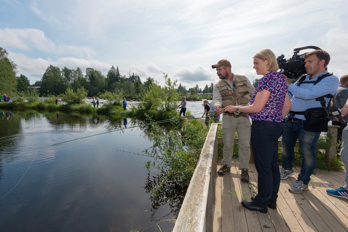 Fluefiskeren Tore Litleré Rydgren (til venstre) instruerer landbruks- og matminister Sylvi Listhaug fra Fremskrittspartiet i fisketeknikk fra en plattform ved Norsk Skogmuseums fiskedam på Prestøya i Glomma.  Rydgren var kledd friluftsantrekk, blant annet med en vest med diverse fiskeutstyr på overkroppen.  Listhaug var kledd i mønstret, kortermet bluse og svart bukse.  Her holder hun fluestanga, og etter hvert fanget hun en liten ørret.  Fjernsynsfotograf Olav T. Hustad Wold fra TV2 fulgte fiskeinstruksjonen med kamera.  Landbruks- og matministeren besøkte jakt- og fiskedagene som foredragsholder i seminaret «Gull og grønne skoger – nye grep for å øke skogens verdi og glede for alle» i museets auditorium.  Etterpå benyttet hun sjansen til å oppleve noe av programmet i museets friluftsmuseum, blant annet her ved fiskedammen. 