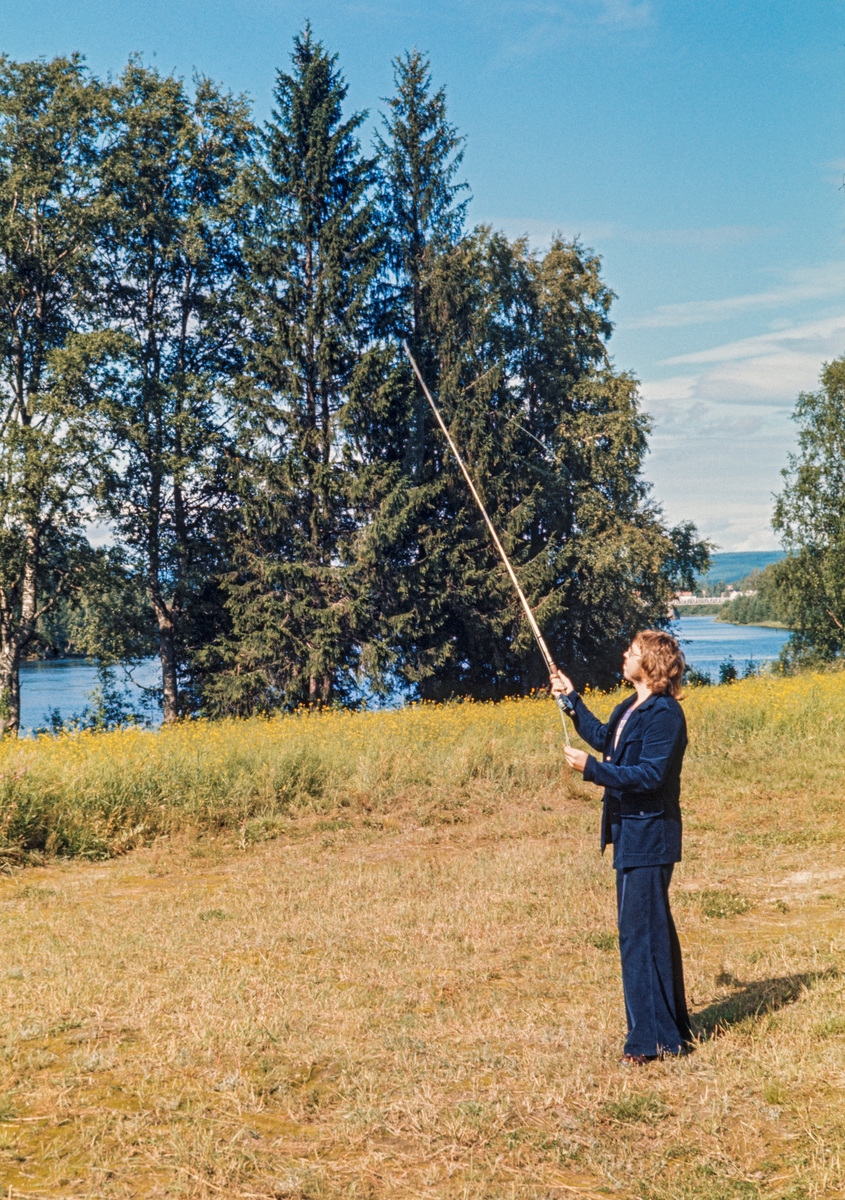 Demonstrasjon av fluekasting under De nordiske jakt- og fiskedager på Norsk Skogbruksmuseum i Elverum i 1973.  Arrangementet ble avviklet 11. og 12. august.  På programmet sto fagforedrag, hundeutstilling, skytekonkurranser, fluebinding, kløvtransport med hund og hest, samt demonstrasjon av fiskeutstyr.  Her ser vi en ung mann med mørkeblå bukse og jakke som kaster med fluestang på grasbevokst mark, vest for den daværende museumsbygningen, med Glomma i bakgrunnen.  Det skal ha vært solgt cirka 3 500 billetter til de to jakt- og fiskedagene i 1973.  Arrangementet gikk parallelt med «Liv i stuene» på Glomdalsmuseet, som også samlet cirka 1 000 museumsgjester. 