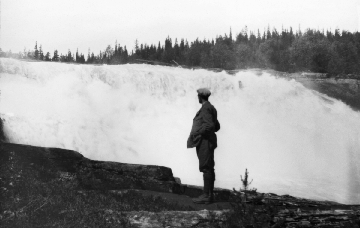 En dresskledd mann med langskaftete lærstøvler og sixpencelue betrakter et stort, kvitskummende fossefall i Mårvassdraget i Tinn i Telemark.  Det er flere slike fall i den nedre delen av vassdraget, blant annet Håkalandsfossen, uten at registrator er lokalkjent nok til å ta stilling til om det er denne fossen vi ser.  Det som er sikkert er at fotografiet er tatt på et tidspunkt da det var mye vann i Mår, som her faller over et bratt stup.  Mannen i forgrunnen betrakter vannmassene fra ei bergflate ved siden av fossen. 