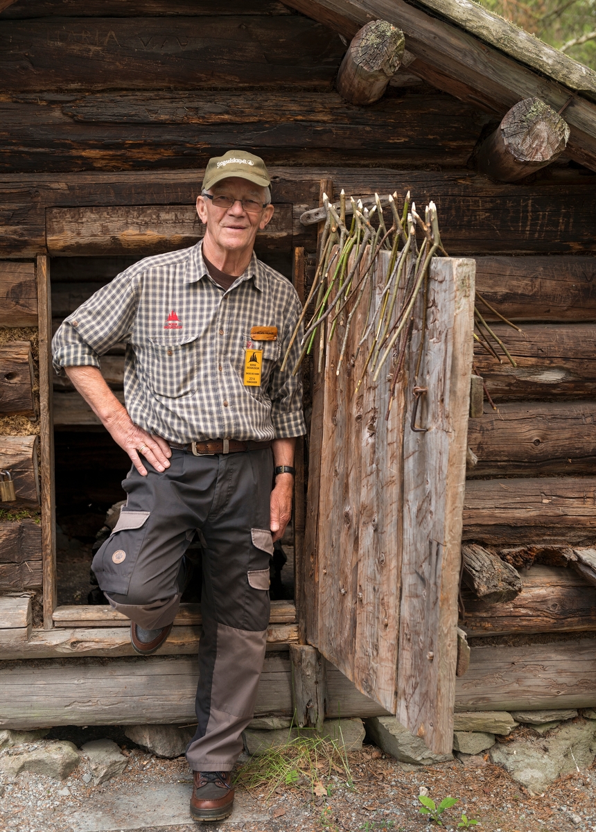 Norsk Skogmuseums mangeårige museumslærer Ingvar Haugen, fotografert i døra til den såkalte «Bjønnbua» fra Rognvola i Stor-Elvdal i museets friluftsmuseum på Prestøya.  Haugen er kledd i det «villmarksantrekket» som i mange år var museets omviserantrekk.  Lua er fra Det norske Skogselskap, og inngikk ikke i denne «uniformen».  På døra ved siden av Ingvar ser vi en masse Y-formete kvister av lauvtrevirke.  Dette har sammenheng med at han da dette fotografiet ble tatt bemannet en av mange «stasjoner» under det store skolerettete arrangementet «Skog og vann 2014».  Ingvar formidlet ønskekvisttradisjonen, og kvistene på dørbladet var naturligvis lagd med henblikk på at elevene sjøl skulle få prøve å gå med ønskekvist, og erfare om de kjente spenninger som kunne tyde på at det for eksempel var ei vannåre i nærheten.  Bruken av ønskekvist er kontroversiell, og fysikere som har gjort tester har benektet at det finnes krefter som virker mellom vannårer og slike kvister som kan forklare utslag i bærerens hender.  De har følgelig karakterisert dette som en «ideomotorisk prosess».  I museet presenteres ønskekvisten som et ledd i et opplegg for tradisjonsformidling.  Dette skjer blant annet under arrangementet «Skog og vann», som arrangeres i begynnelsen av juni hvert år.  Ei kort orientering om dette arrangementet finnes under fanen «Opplysninger». 