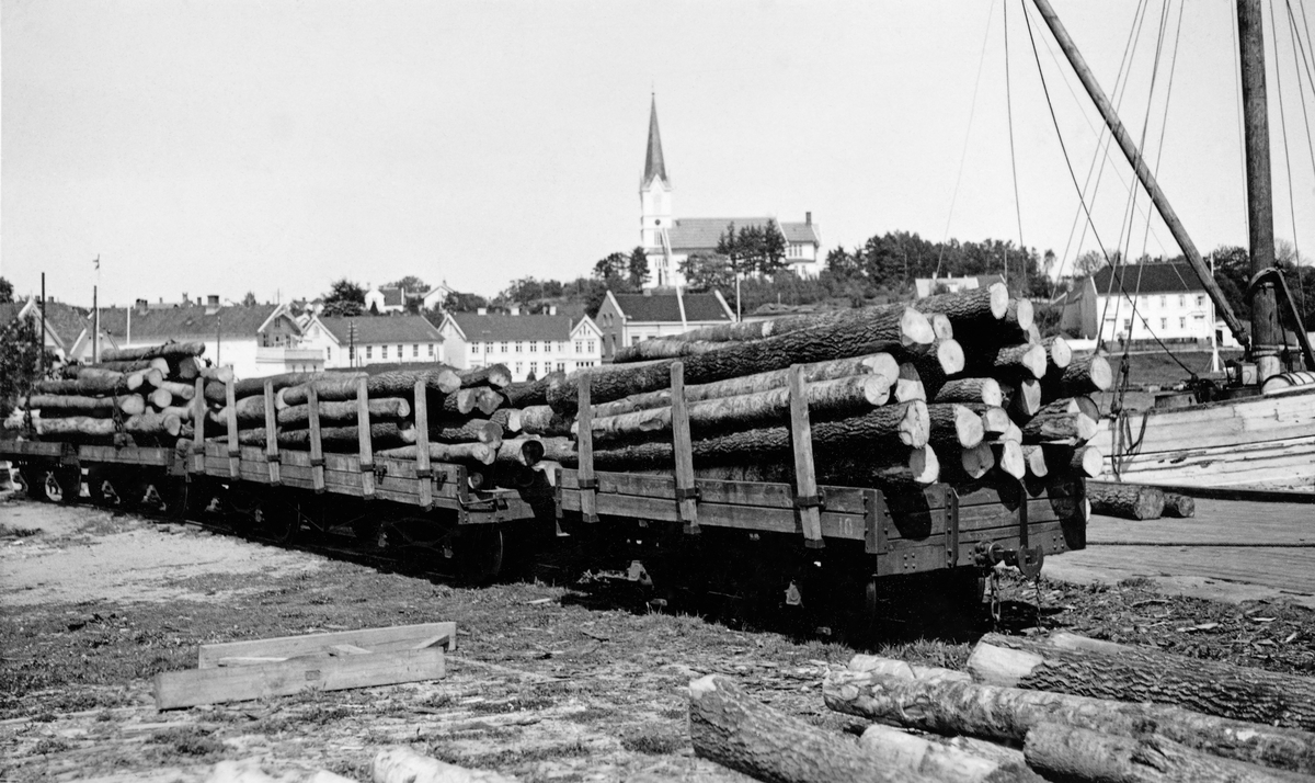 Jernbanevogner med ospetømmer på jernbanespor ved kaia i kystbyen Lillesand i Aust-Agder.  Fotografiet skal være tatt i 1935.  Vi ser tre åpne jernbanevogner med ubarket ospetømmer.  I forgrunnen ligger det også noen stokker på bakken.  Bildet er tatt i forbindelse med omlasting.  Tømmeret skulle transporteres videre med båt til fyrstikkfabrikken på Agnes ved Stavern i Vestfold.  I bakgrunnen skimter vi de kvitmalte husene i havneområdet i Lillesand, og på bakkekammen bakenfor ligger Lillesand kirke, ei tidstypisk trekirke fra slutten av 1800-tallet.

Sommeren 2024 fikk museet denne utdypende kommentaren til fotografiet fra Kari Michelsen:

«Treslaget osp ble også i stor grad benyttet ved produksjon av treull. Sørlandets treullfabrikk (1922-60) i Lillesand var en av de få slike fabrikker i Norge.»