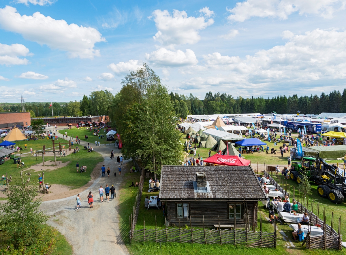 Utsikt fra balkongen utenfor oppholdsrommet i det rekonstruerte brannvakttårnet på Norsk Skogmuseum i Elverum.  Fotografiet er tatt mot gangvegen mellom museumsbygningen og friluftsmuseet på Prestøya søndag 10. august 2014, den siste dagen under De nordiske jakt- og fiskedager dette året, noe som betydde at det var flere mennesker og mer aktivitet på uteområdet enn på en vanlig søndag.  Til høyre i forgrunnen ser vi skogvokterboligen som er flyttet til museet fra Galten i Engerdal.  Ved dette huset var det serveringssted med salg av kaffe, vafler og is under arrangementet.  Til venstre i forgrunnen ser vi utkanten av lekeområdet for barn o bakenfor ser vi lavvoen der den statlige skogforvaltningen presenterte sin virksomhet for publikum.  På motsatt side av gangvegen ser vi det såkalte «håndverkstorget», til venstre for trerekka som står i grensesonen mellom det som for noen tiår siden var skillet mellom to åkrer. På sørsida av denne ser vi det såkalte «frilandsområdet», der de fleste av arrangementets kommersielle aktører var samlet.  Lassbæreren til høyre i forgrunnen står der som et landemerke som markerer det såkalte «skogbrukstunet» på bakkekammen bak våningshuset på klokkergarden Fossum, der organisasjonene i skogbruket hadde sine aktiviteter under arrangementet.  Den dagen da dette fotografiet ble tatt var det 6 789 personer som løste billetter til De nordiske jakt- og fiskedager.