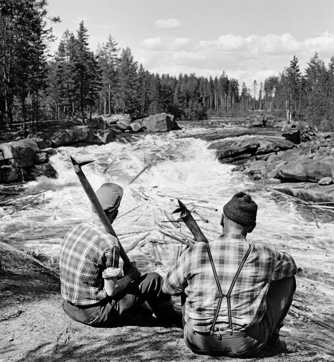 To tømmerfløtere, utstyrt med fløterhaker og kledd i rutete bomullsskjorter, fotografert sittende på en bergnabb nedenfor en foss i den øvre delen av Flisavassdraget, antakelig ett av de såkalte Flisfallene.  Bak karene, som satt med ryggen mot fotografen, ser vi hvordan elva faller i en kvitskummende foss over en bergrygg, omgitt av furuskog.  I det kvitskummende elvevatnet ser vi en del tømmerstokker.  Bildet er tatt i 1969. Fløtere. Fløting av tømmer. Skogbruk.