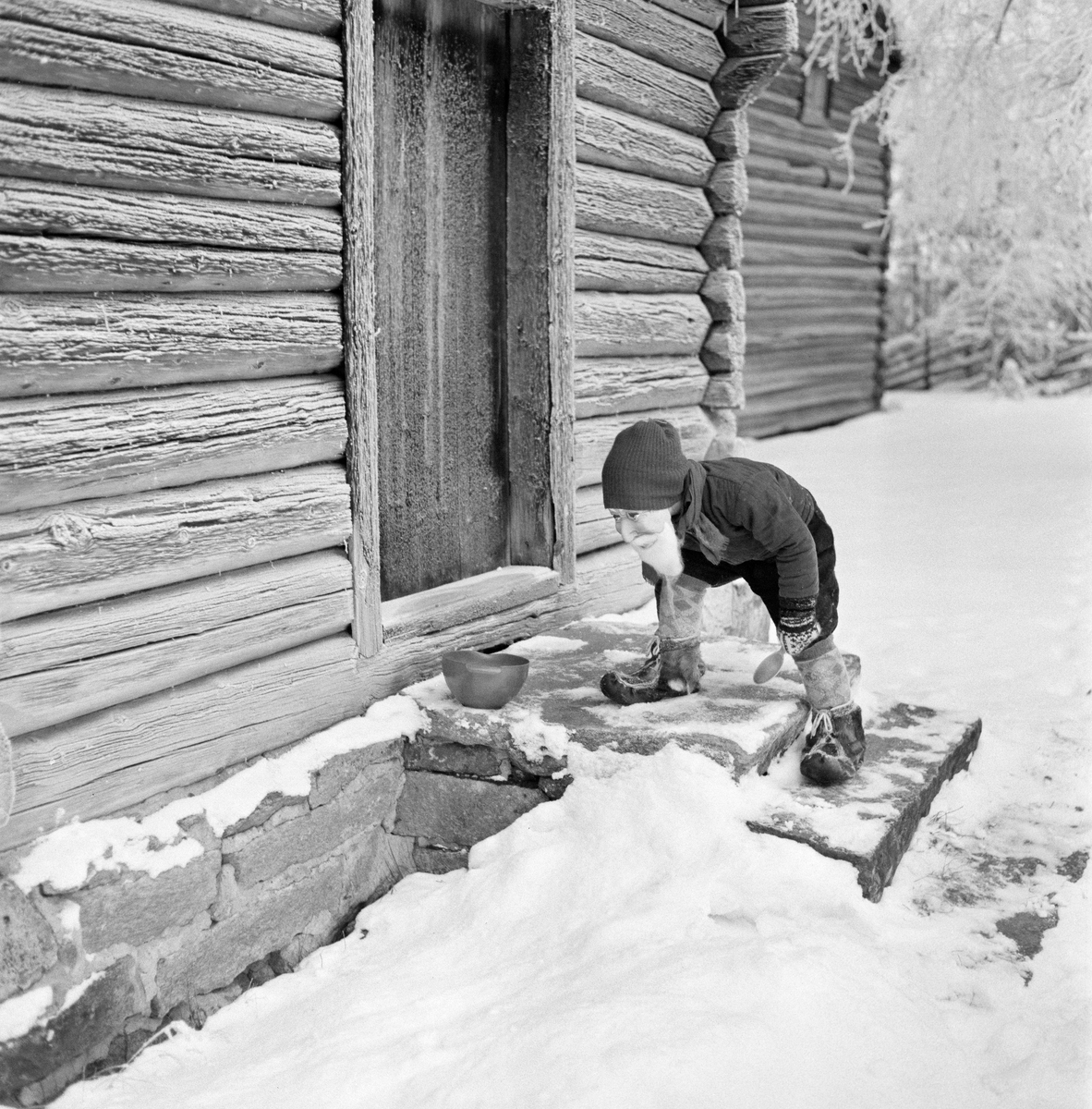 Nisseskikkelse på trammen framfor barfrøstua fra Bjønngarden (Bjøntegård) i Rendalen, slik den står i Glomdalsmuseets friluftsmuseum i Elverum.  Fotografiet er tatt en kald vinterdag før jul i 1963.  Marka var snødekt, og de sprukne lafteveggene på barfrøstua og det bakenforstående Høye-loftet var dekt av rim. Nisseskikkelsen sto på trappa, med ei stor skje i den ene handa og bøyde seg mot en grøtbolle som var plassert på dørhella, like inntil inngangsdøra.