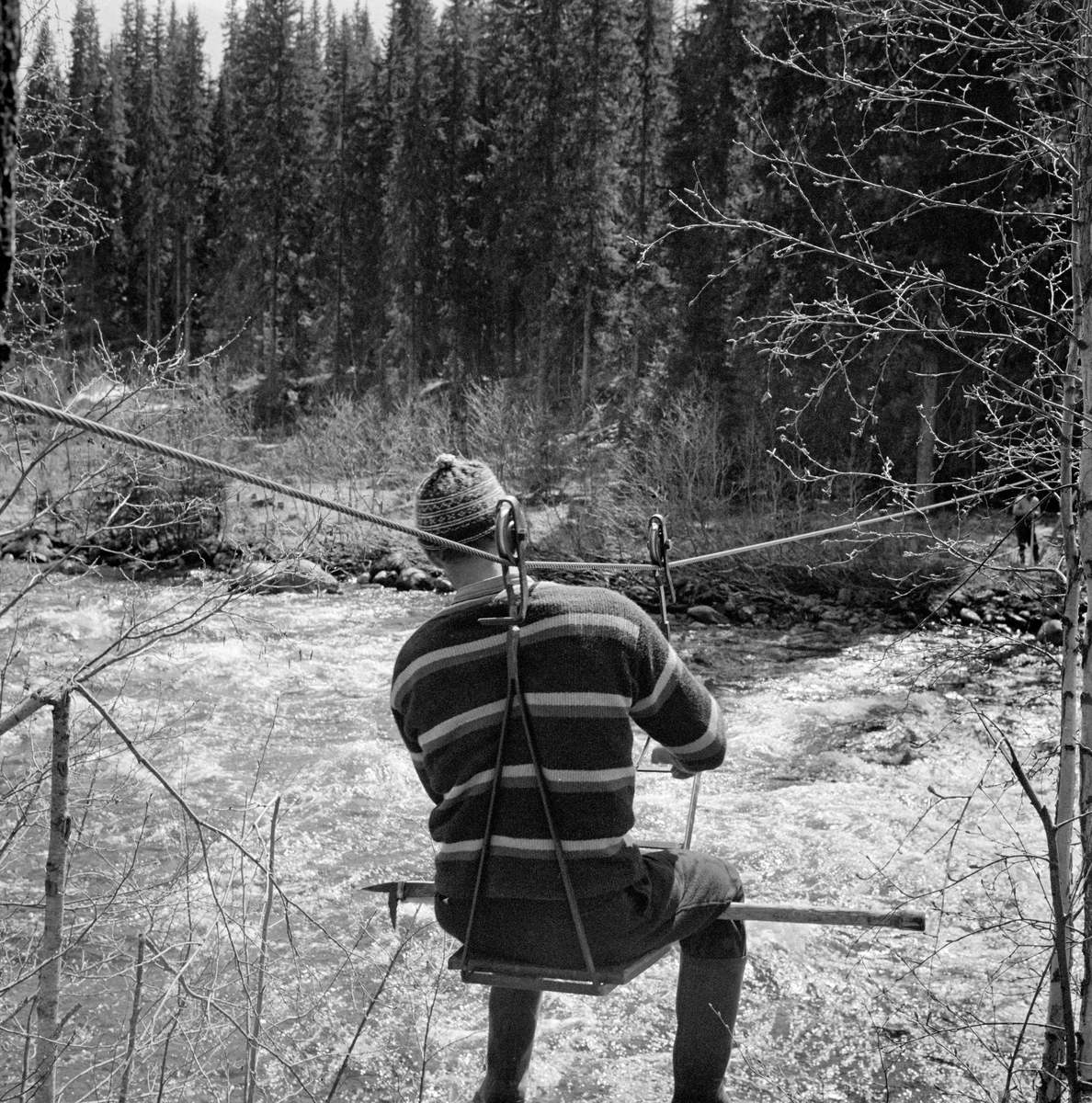 Tømmerfløter, fotografert under kryssing av elva Åsta på fløterstol våren 1969. Denne innretningen er lagd av to buete stålstenger som møtes i ytterendene, der det er festet trinser, som ruller på en stålvaier som er spent på tvers av elveløpet. Fløteren sitter på ei setefjøl med fløterhaken på tvers under lårene og med den nevnte vaieren (bærekabelen) i hodehøyde. Han beveger stolen – og dermed seg sjøl – på tvers av elveløpet ved å gripe rundt vaieren og trekke seg fram. Fløteren var kledd i mørke bukser og stripete ullgenser. Han hadde støvler på beina og topplue på hodet.  I bakgrunnen ser vi hvordan vaieren var forankret i en trestamme, støttet av ei A-formet treramme. Fløterstoler av denne typen ble installert på steder langs fløtingsvassdragene der tømmeret hadde lett for å sette seg fast («binde seg»), og der det var vanskelig både å krysse vassdraget og å komme seg ut til grusører eller steinskjær der det hadde lett for å danne seg vaser (hauger) av sammenstuvete tømmerstokker. Fløterstoler ble også mye brukt ved vassdrag der isgang kunne vanskeliggjøre kryssing i vårsesongen. Oppfinneren, landhandler Eberhard Kokkin (1856-1920), bodde sjøl ved den nedre delen av Åsta. Det er grunn til å anta at han må ha fått idéen til fløterstolen ved å observere utfordringene fløterne i denne elva møtte i sin arbeidshverdag. I 1960-åra var det spent 30 vaiere over Åsta, der fløterne kunne krysse elva på slike fløterstoler.