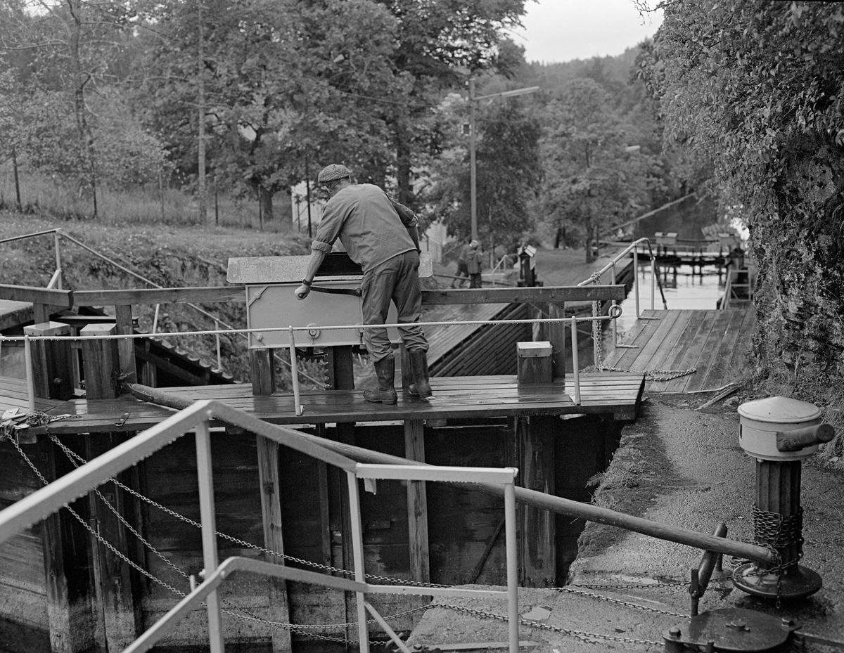 Slusing av tømmer ved Ørje i Marker kommune i Østfold høsten 1982.  Sluseanlegget her betjenes manuelt.  I forgrunnen til høyre ser vi gangspillet som brukes når sluseporten skal åpnes eller stenges.  Ved lukking er det trestanga som går fra gangspillet til porten som skyver den på plass.  Da dette fotografiet ble tatt var den lukket, men Kåre Granli (1921-1991) var i ferd med å åpne luka nederst på porten, slik at mye av vannet kunne strømme over i nedenforliggende slusekammer.  Dette ble gjort ved hjelp av ei sveiv på jernkassa som skjermet tannhjulet og den øvre delen av tannstanga som justerte lukas posisjon.  Når nedenforliggende slusekammer var fylt av vann fra det ovenforliggende kunne portene åpnes, slik at tømmeret kunne flyte over.  På denne måten flyttet man slusevendingene – i dette tilfellet fire bunter massevirke – trinn for trinn gjennom tre slusekamre fra Rødenessjøen ovenfor 10 høydemeter ned til Ørjeelva nedenfor.

En liten historikk om tømmerfløting og kanaliseringsarbeid i Haldenvassdraget finnes under fanen «Opplysninger».