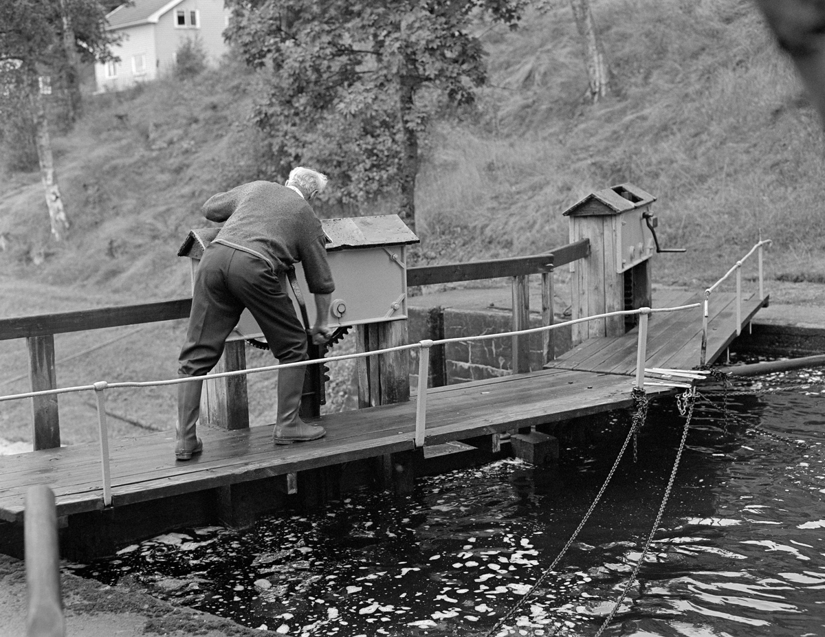 Fra sluseanlegget ved Ørje i Marker kommune i Østfold.  Fotografiet er tatt i 1982, som var den siste sesongen det ble fløtet tømmer på Haldenvassdraget.  Vi ser slusemester Odd Johansen (1917-1993) idet han åpner lukene på en av sluseportene.  Dette ble gjort ved hjelp av ei sveiv, som ble plassert på en tapp på ei jernbeslått kasse som var montert ved rekkverket på sluseporten.  Den nevnte jernkassa skjermet tannhjulet og den øvre delen av tannstanga som åpnet eller lukket luka på den nedre delen av porten.  Ved åpning av slike luker strømmet det vann over i nedenforliggende slusekammer.  Ved tømmerslusing fikk denne vannstrømmen slusevendinga til å drive framover mot neste kammer.  Tappinga via lukene utliknet også vannstanden i ovenforliggende og nedenforliggende slusekammer, slik at portene kunne åpnes og slusegodset (tømmer eller båter) kunne beveges i riktig retning.  Odd Johansen var i mange år slusemester ved Strømsfoss i Aremark, lengre nede i vassdraget.  I 1982 var Johansen pensjonist og kunne dermed kunne han også hjelpe til med fløtinga andre steder i vassdraget i en situasjon der det, fordi Haldenvassdragets Fellesfløtingsforening skulle avvikles, var litt knapt med mannskap i yrkesaktiv alder.

En liten historikk om tømmerfløting og kanaliseringsarbeid i Haldenvassdraget finnes under fanen «Opplysninger».