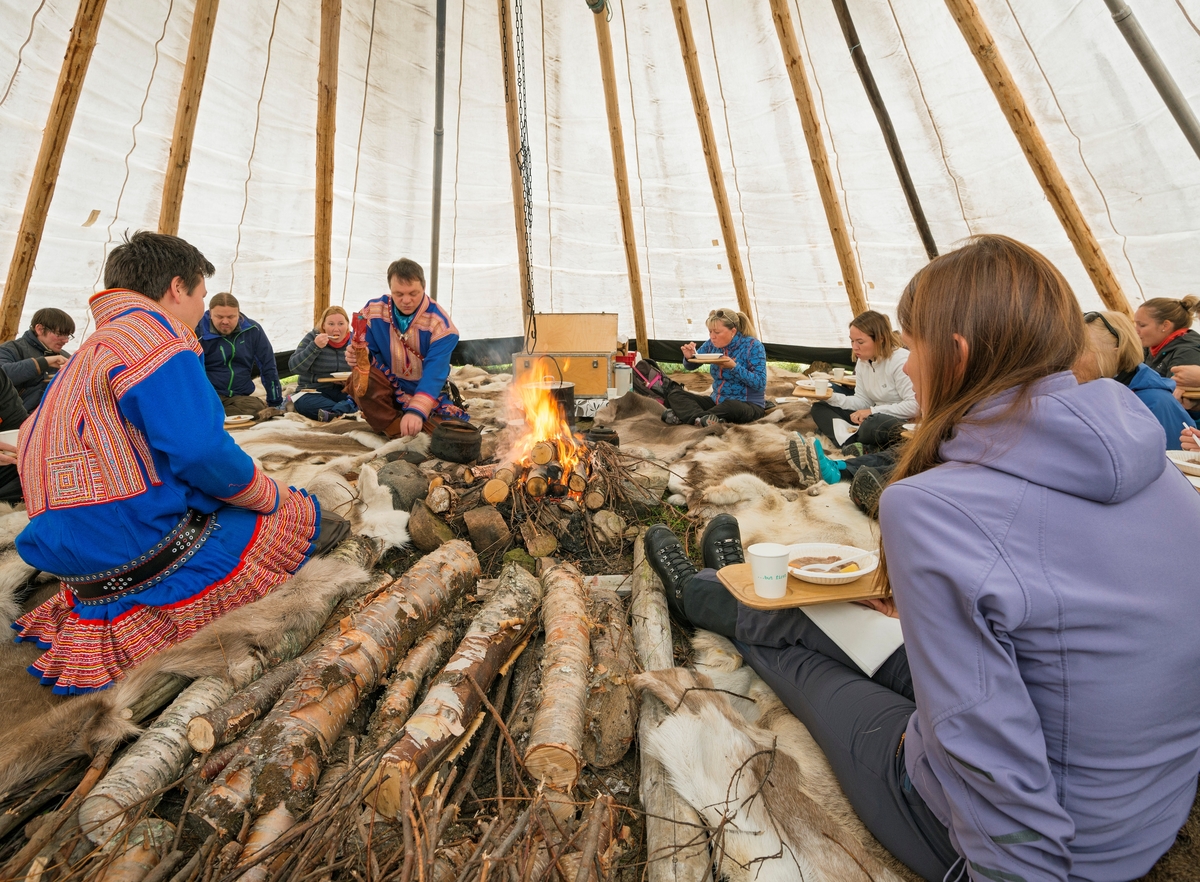 Delegasjon fra Norges vassdrags- og energidirektorats (NVEs) museumsordning samlet i en lavvo i Masi (Máze) i Finnmark.  Lavvoen tilhører Johan P. Eira og hans firma, Cavzo Safari, som tilbyr turistopplevelser i det lokale landskapet, med blant annet denne lavvoen og elvebåter som arenaer, og med vekt på de samiske tradisjonene i området.  Da dette fotografiet ble tatt serverte vertskapet, Johan P. Eira og sønnen hans, mat til gjestene, som satt på reinsdyrskinn rundt den åpne varmen i midten av lavvoen.