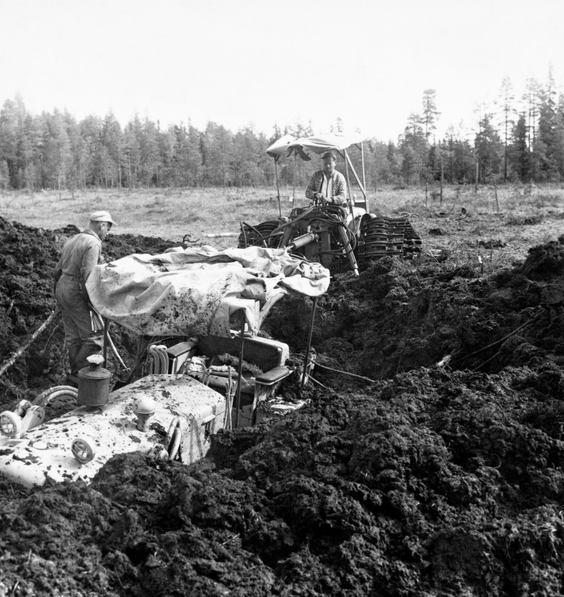 Beltetraktor, djupt nedsunket i myr på Grue Finnskog i juni 1961.  Uhellet skjedde i forbindelse med grøfteforsøk der den store, tunge, finskproduserte «Lokomo-plogen» ble testet.  Dette var ikke første gang at traktorene sank ned i myrtorva under slike forsøk.  Da forsøkene startet fire år tidligere brukte man 12 tonn tunge beltetraktorer, slik finnene gjorde, men det viste seg at de sank ned i de våte norske myrene.  Forsøksleder Ivar Samset og hans mannskap gikk derfor over til å bruke noe lettere beltetraktorer, der beltene ble påmontert 85 centimeter lange treelementer, som skulle gi bedre bæreevne.  Likevel hendte det at traktorene sank ned i den våte myrtorva, som da dette fotografiet ble tatt.  Her måtte det leies inn maskiner for å få dem kostbare beltetraktoren opp igjen, noe som i Det norske Skogforsøksvesens album er beskrevet slik: «Opptauing av nedsunket bulldozer. Grøfteforsøkene.  Falken tok 7 500,- kr. for arbeidet.  Vi leide i tillegg bulldozer fra Glomma fellesfløtningsforening (kr. 500) og holdt 5 mann.  Falken er dyr og kan ikke anbefales.»  Mer informasjon om Lokomo-plogen finnes under fanen «Opplysninger».