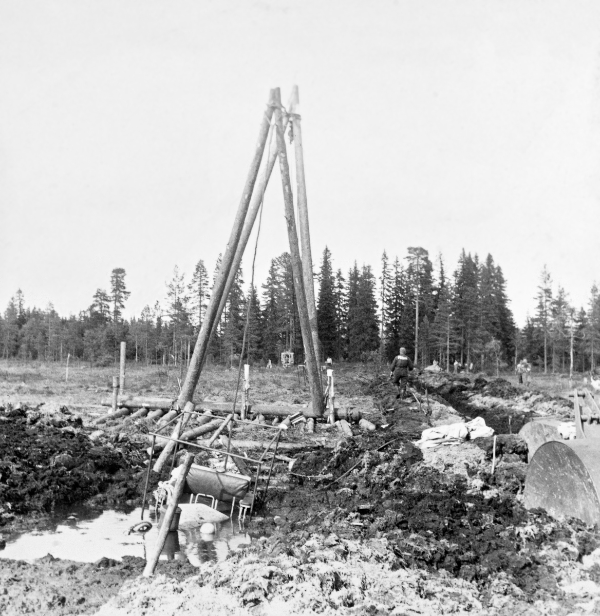 Berging av beltetraktor som hadde sunket djupt ned i ei myr på Grue Finnskog i juni 1961.  Den havarerte traktoren lå til venstre for dette bildeutsnittet.  Fotografiet viser en tømmerflåte på myra, der det sto to omvendt V-formete bukker.  Vaieren fra den havarerte traktoren (til venstre) til kjøretøyene som skulle få den opp (til høyre) gikk via toppen på disse bukkene, antakelig for å for å få trukket den opp fra de våte torvmassene, ikke horisontalt bortover i myra.  Uhellet skjedde i forbindelse med grøfteforsøk der den store, tunge, finskproduserte «Lokomo-plogen» ble testet.  Dette var ikke første gang at traktorene sank ned i myrtorva under slike forsøk.  Da forsøkene startet fire år tidligere brukte man 12 tonn tunge beltetraktorer, slik finnene gjorde, men det viste seg at de sank ned i de våte norske myrene.  Forsøksleder Ivar Samset og hans mannskap gikk derfor over til å bruke noe lettere beltetraktorer, der beltene ble påmontert 85 centimeter lange treelementer, som skulle gi bedre bæreevne.  Likevel hendte det at traktorene sank ned i den våte myrtorva, som da dette fotografiet ble tatt.  Her måtte det leies inn maskiner for å få dem kostbare beltetraktoren opp igjen, noe som i Det norske Skogforsøksvesens album er beskrevet slik: «Opptauing av nedsunket bulldozer. Grøfteforsøkene.  Falken tok 7 500,- kr. for arbeidet.  Vi leide i tillegg bulldozer fra Glomma fellesfløtningsforening (kr. 500) og holdt 5 mann.  Falken er dyr og kan ikke anbefales.»  Mer informasjon om Lokomo-plogen finnes under fanen «Opplysninger».