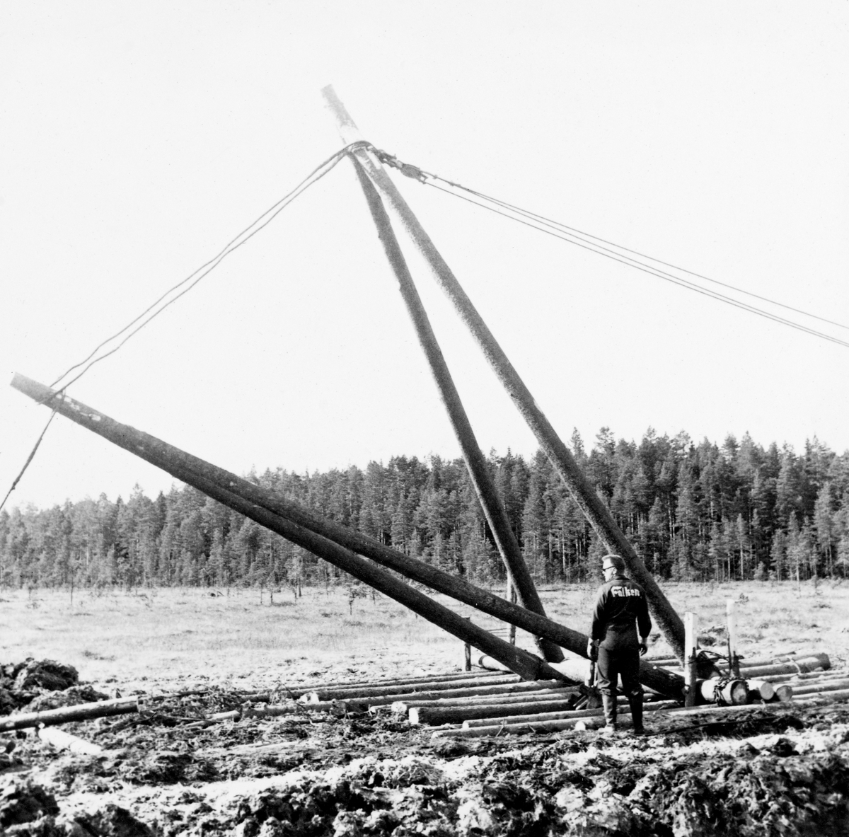 Berging av beltetraktor som hadde sunket djupt ned i ei myr på Grue Finnskog i juni 1961.  Den havarerte traktoren lå til venstre for dette bildeutsnittet.  Fotografiet viser en tømmerflåte på myra, der det sto to omvendt V-formete bukker.  Vaieren fra den havarerte traktoren (til venstre) til kjøretøyene som skulle få den opp (til høyre) gikk via toppen på disse bukkene, antakelig for å for å få trukket den opp fra de våte torvmassene, ikke horisontalt bortover i myra.  Uhellet skjedde i forbindelse med grøfteforsøk der den store, tunge, finskproduserte «Lokomo-plogen» ble testet.  Dette var ikke første gang at traktorene sank ned i myrtorva under slike forsøk.  Da forsøkene startet fire år tidligere brukte man 12 tonn tunge beltetraktorer, slik finnene gjorde, men det viste seg at de sank ned i de våte norske myrene.  Forsøksleder Ivar Samset og hans mannskap gikk derfor over til å bruke noe lettere beltetraktorer, der beltene ble påmontert 85 centimeter lange treelementer, som skulle gi bedre bæreevne.  Likevel hendte det at traktorene sank ned i den våte myrtorva, som da dette fotografiet ble tatt.  Her måtte det leies inn maskiner for å få dem kostbare beltetraktoren opp igjen, noe som i Det norske Skogforsøksvesens album er beskrevet slik: «Opptauing av nedsunket bulldozer. Grøfteforsøkene.  Falken tok 7 500,- kr. for arbeidet.  Vi leide i tillegg bulldozer fra Glomma fellesfløtningsforening (kr. 500) og holdt 5 mann.  Falken er dyr og kan ikke anbefales.»  Mer informasjon om Lokomo-plogen finnes under fanen «Opplysninger».