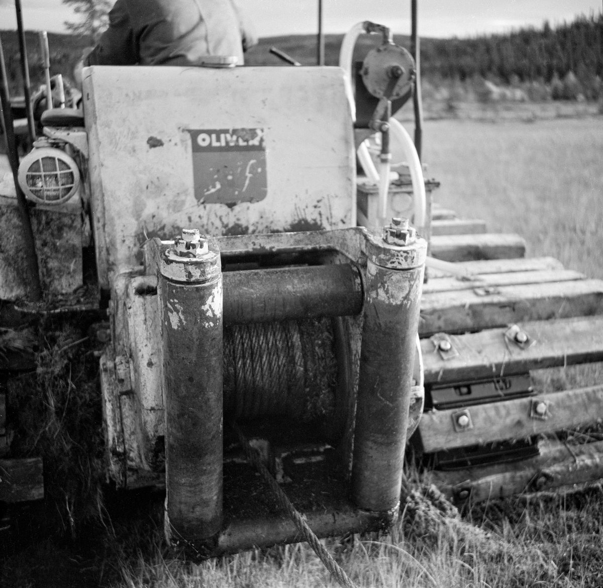 Bakmontert, entromlet vinsj på beltetraktor (bulldoser) av merket Oliver.  Fotografiet er tatt under grøfteforsøk med såkalt «Lokomo-plog» i Trysil høsten 1961.  Plogen som beltetraktoren og vinsjen skulle trekke gjennom fuktig myrtorv veide 3,8 tonn.  Den etterlot seg ei grøftefure som var 70-90 centimeter djup, 30 centimeter bred i botnen og en drøy meter bred i toppen.  En rullekniv skar et snitt i torva framfor plogbrynet.  Vridde veltefjøler av stål, med forlengelser av tre, sørget for at torva ble løftet opp og skjøvet til side.  Dette krevde naturligvis stor kraft.  Pløyinga foregikk etappevis – traktoren ble kjørt 40-60 meter framover i den retningen en ønsket at grøfta skulle gå.  Deretter ble plogen vinsjet etter, inntil den sto 10-15 meter bak traktoren, som deretter ble flyttet nye 40-60 meter framover.  Slik fortsatte arbeidet.  Det ble montert 85 centimeter lange fjøler på traktorbeltene for å forebygge at beltetraktoren sank ned i blautmyr når trykket ble for stort.