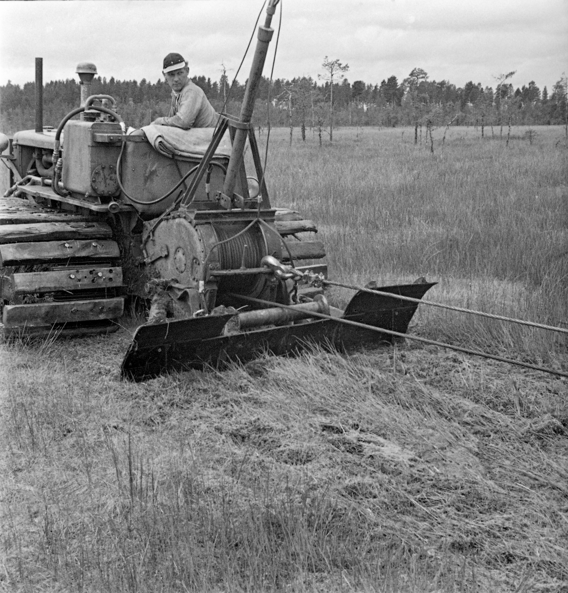 Beltetraktor med bakmontert vinsj og skjær (anker), brukt under forsøk med den finskproduserte «Lokomo-plogen» på Grue Finnskog i 1957.  Denne plogen ble ikke koplet direkte på traktoren, men i stedet trukket gjennom myra bak dette kjøretøyet ved hjelp av en vinsj mens traktoren sto stille, med det nevnte skjæret eller ankeret presset ned mot bakken.  Fotografiet viser også hvordan karene som arbeidet med disse forsøkene hadde montert planker på stålbeltene for å gjøre dem bredere og dermed gi kjøretøyet bedre bæreevne.  Lokomo-plogen etterlot seg ei grøftefure som var 70-90 centimeter djup, 30 centimeter bred i botnen og en drøy meter bred i toppen.  En rullekniv skar et snitt i torva framfor plogbrynet.  Vridde veltefjøler av stål, med forlengelser av tre, sørget for at torva ble løftet opp og skjøvet til side.  Dette krevde naturligvis stor kraft.  Pløyinga foregikk etappevis – traktoren ble kjørt 40-60 meter framover i den retningen en ønsket at grøfta skulle gå.  Deretter ble plogen vinsjet etter, inntil den sto 10-15 meter bak traktoren, som deretter ble flyttet nye 40-60 meter framover.  Slik fortsatte grøftinga, flytt for flytt.  Da dette fotografiet ble tatt var det Kåre Andersen som satt på traktoren.  Han var ansatt som teknisk assistent ved Skogforsøksvesenets driftstekniske avdeling. Mer informasjon om Lokomo-plogen finnes under fanen «Opplysninger».