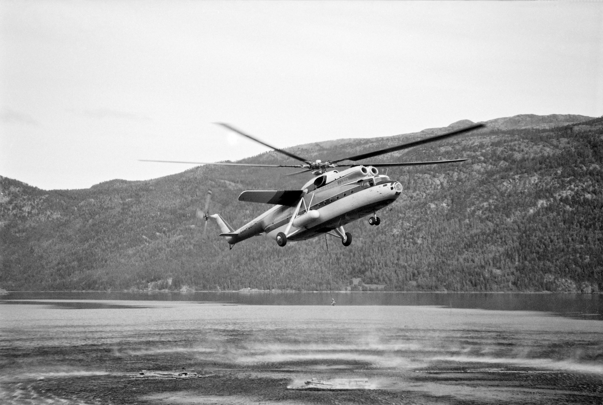 Helikopter av typen MI-6, fotografert under nedstigning mot Kviteseidvatnet i Telemark under forsøk med tømmertransport fra det bratte fjellterrenget sør for Dyreskarnut ned til fløtingsvassdraget.  Fotografiet er tatt idet helikopterpiloten nettopp hadde sluppet en tømmerbunt ned på Kviteseidvatnet.  Den avbildete maskinen var i midten av 1960-åra verdens største helikopter.  Helikopterkroppen var 33,2 meter lang og rotorbladene spente over en diameter på 35 meter.  Maskinen veide 40,5 tonn og kunne i tillegg frakte 12 tonn nyttelast i et romslig, men enkelt utstyrt lasterom.  MI-6 hadde to kraftige gassturbinmotorer, som til sammen kunne utvikle 11 000 hestekrefter med et drivstofforbruk på 3 500 – 4 000 liter i timen.  I Kviteseid ble maskinen testet på henting av buntet tømmer fra den nevnte lokaliteten sør for Dyrskarnut, 4 860 meter fra og 390 meter høyere enn slippstedet i Kviteseidvatnet.  Tømmeret på hogstfeltet var på forhånd samlet i bunter med drøyt 5 tonns vekt og gjennomsnittlig 8,5 kubikkmeters volum (fastmasse).  Buntene ble hengt under helikopteret i cirka 10 meter lange bærestropper.  Konklusjonen fra disse undersøkelsene ble at kostnadsbildet minte om det man hadde hatt da man et par år tidligere hentet noe tømmer fra en annen lokalitet i området med et noe mindre helikopter et par år tidligere.  Den store russiske maskinen kunne riktignok ta noe større bunter, men på kløftet, blokkrik mark som her innebar det tidkrevende manuelt slit å samle store tømmerbunter.  «Selv om selve helikoptertransporten blir noe billigere med en større maskin, vil de øvrige kostnader bli noe høyere», skrev forsøkslederen, professor Ivar Samset (1918-2015).  Det han ikke skrev, men erfarte, var at helikoptertransport av tømmer var tapsbringende.  Driftskostnadene knyttet til arbeidet på hogstfeltet og bruken av helikopteret oversteg tømmerets omsetningsverdi.  Mer informasjon om forsøkene med helikoptertransport av tømmer fra vanskelig terreng finnes under fanen «Opplysninger».