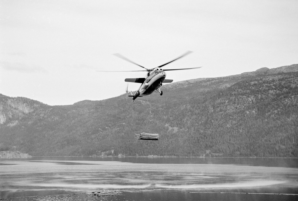 Helikopter av typen MI-6, fotografert under nedstigning mot Kviteseidvatnet i Telemark under forsøk med tømmertransport fra det bratte fjellterrenget sør for Dyreskarnut ned til fløtingsvassdraget.  Fotografiet ble tatt idet helikopteret nærmet seg en høyde over vannspeilet der tømmerbunten – «hivet» – kunne slippes uten nevneverdig fare for skade på tømmeret.  MI-6 var i midten av 1960-åra verdens største helikopter.  Helikopterkroppen var 33,2 meter lang og rotorbladene spente over en diameter på 35 meter.  Maskinen veide 40,5 tonn og kunne i tillegg frakte 12 tonn nyttelast i et romslig, men enkelt utstyrt lasterom.  MI-6 hadde to kraftige gassturbinmotorer, som til sammen kunne utvikle 11 000 hestekrefter med et drivstofforbruk på 3 500 – 4 000 liter i timen.  I Kviteseid ble maskinen testet på henting av buntet tømmer fra den nevnte lokaliteten sør for Dyrskarnut, 4 860 meter fra og 390 meter høyere enn slippstedet i Kviteseidvatnet.  Tømmeret på hogstfeltet var på forhånd samlet i bunter med drøyt 5 tonns vekt og gjennomsnittlig 8,5 kubikkmeters volum (fastmasse).  Buntene ble hengt under helikopteret i cirka 10 meter lange bærestropper.  Konklusjonen fra disse undersøkelsene ble at kostnadsbildet minte om det man hadde hatt da man et par år tidligere hentet noe tømmer fra en annen lokalitet i området med et noe mindre helikopter et par år tidligere.  Den store russiske maskinen kunne riktignok ta noe større bunter, men på kløftet, blokkrik mark som her innebar det tidkrevende manuelt slit å samle store tømmerbunter.  «Selv om selve helikoptertransporten blir noe billigere med en større maskin, vil de øvrige kostnader bli noe høyere», skrev forsøkslederen, professor Ivar Samset (1918-2015).  Det han ikke skrev, men erfarte, var at helikoptertransport av tømmer var tapsbringende.  Driftskostnadene knyttet til arbeidet på hogstfeltet og bruken av helikopteret oversteg tømmerets omsetningsverdi.  Mer informasjon om forsøkene med helikoptertransport av tømmer fra vanskelig terreng finnes under fanen «Opplysninger».