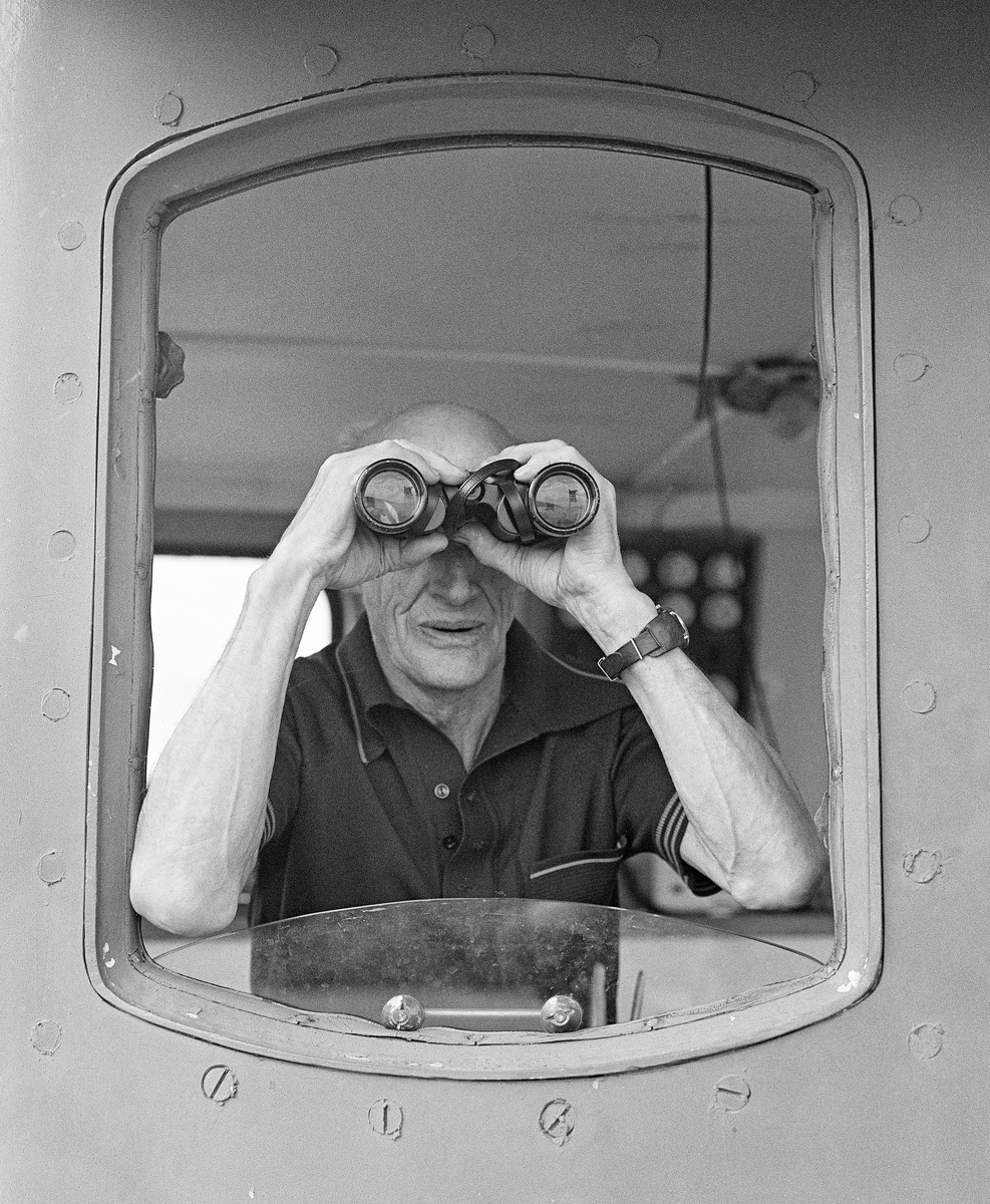Rendølen Sigvart Svalstad (1915-2003), fotografert mens han speidet ut av et av vinduene i styrhuset på slepebåten «Storsjø» på innsjøen med samme navn i Ytre Rendalen i Hedmark.  Kikkerten var et nyttig hjelpemiddel, både når en skulle se at ringbommen bak båten var intakt, og når man, som her arbeidet med sluttrensk og skulle ha med seg alle restbeholdninger som lå og fløt langs strendene.  Sigvart Svalstad ble tatt inn i besetningen på slepebåten Storsjø i 1942, da under ledelse av sesongansatte sjøfolk fra Østfold.  Etter at dampmaskinen ble erstattet av en dieselmotor i 1957-58, var det lokale folk under ledelse av veteranen Halvor Fiskvik (1908-1989).  Etter at Fiskvik ble pensjonist fikk Svalstad gleden av å være skipper de siste tre sesongene det foregikk tømmerfløting på Storsjøen.  Båten hadde da en besetning på fire mann, skipperen inkludert.