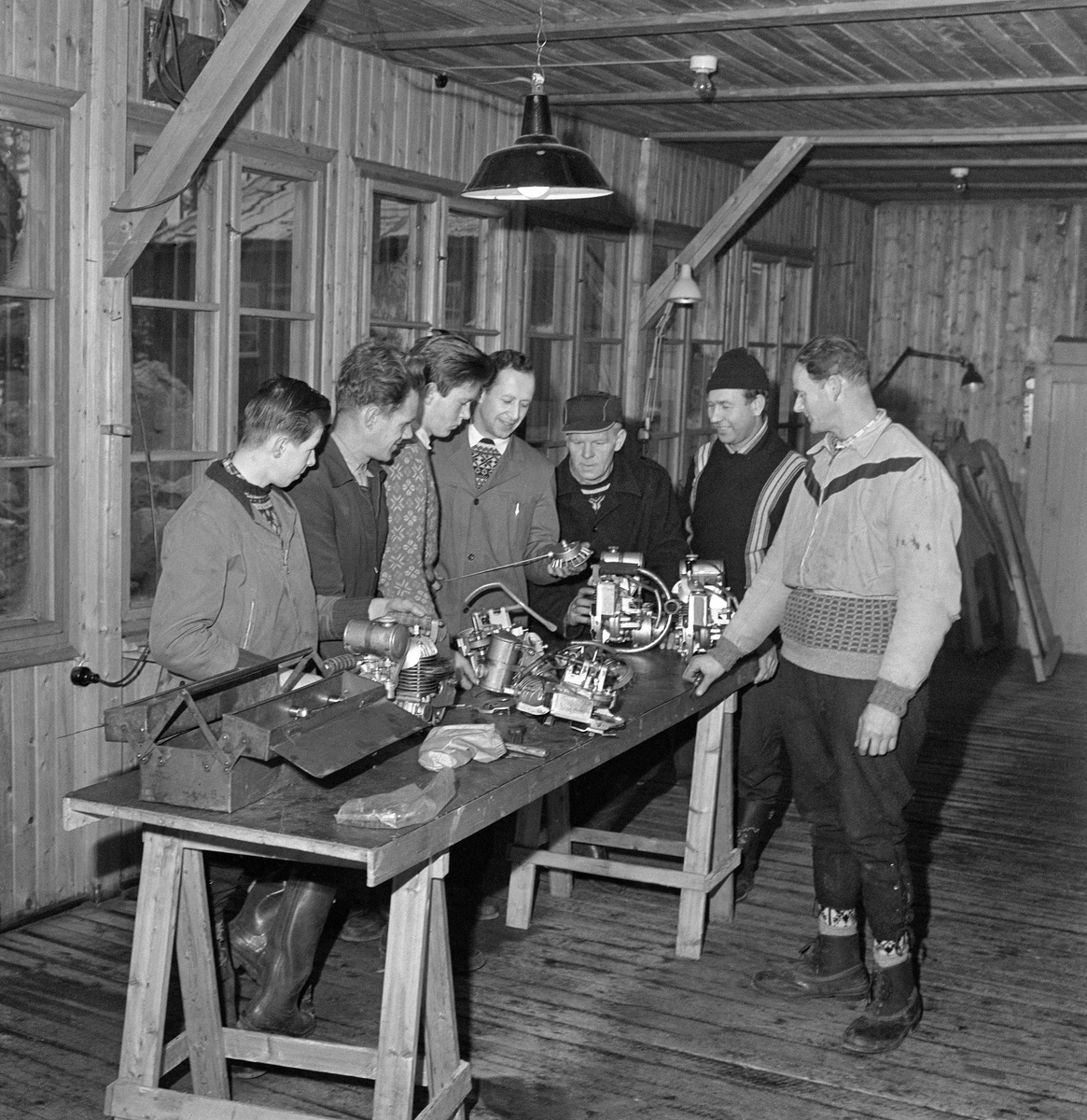 Instruktør Gangnes fra motorsagfabrikken Jobu orienterer skogsarbeidere ved yrkesskolen på Sønsterud i Åsnes i Solør i motorsagas mekanikk.  Undervisninga foregikk ved et bord som sto på to bukker.  På den ene enden av bordplata sto det ei redskapskasse av stål.  Ellers var bordet dekt av komponenter fra to motorsager, som man hadde skrudd fra hverandre.  Nærmest redskapskassa (til venstre på bordet) ser vi komponenter fra en Jobu junior, til høyre ser vi en Comet.  Da fotografiet ble tatt sto Gangnes med et viftehus og ei startsnor i hendene, og de seks kursdeltakerne fulgte interessert med.  Skogsarbeiderskolen på Sønsterud ble etablert i 1946.  Den tilbød kurs av varierende lengde, tolv uker for nybegynnere og 4-5 uker for mer vante folk.  Skolen tilbød også spesialkurs med temaer som motorsag, vinsj og transportteknikk.  De første årene foregikk undervisningen hovedsakelig i en del brakker som sto igjen etter den såkalte «Arbeidstjenesten», som hadde mye aktivitet i dette området under 2. verdenskrig.  Den motorsagtekniske undervisningen på dette bildet foregikk i ei av brakkene.  At slike emner var så sentrale i det pedagogiske tilbudet avspeilet den store moderniseringa av skogsarbeidet etter 2. verdenskrig, med overgang fra handredskaper til motorisert materiell.  De nye redskapene var kostbare i innkjøp.  Derfor ble det oppfattet som viktig skogsarbeiderne visste å handtere motorsager og annet mekanisk materiell på en effektiv måte, og at de var sjølhjulpne dersom det oppsto driftsstans, slik at uproduktive avbrudd ble kortest mulig.  Dette fotografiet ble antakelig tatt i 1960.  Da var Kirke- og undervisningsdepartementet i ferd med å reise et mer moderne undervisnings- og internatbygninger.  Dette var sannsynligvis den siste sesongen den motorsagtekniske undervisningen foregikk i denne brakka.