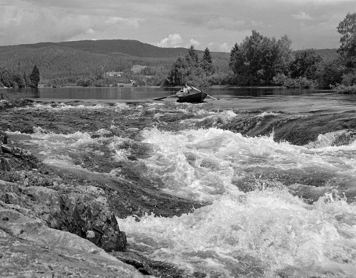 Sluttrensk i Norsfossen i Glomma i begynnelsen av juli 1985.  Fotografiet viser fløteren Sigurd Sondre Huvestad, som var i ferd med å ro fra en stilleflytende del av elva, mot et mer opprørt stryk, der vannet gikk kvitskummende.  Mannen var en tømmerfløter i en av Glomma fellesfløtingsforenings robåter.  I bakgrunnen ser vi mot det skogkledde Vålerberget med et toppunkt 310 meter over havet.  Sjølve Norsfossen ligger cirka 150 meter over havet.  1985 var den siste sesongen med ordinær tømmerfløting i Glomma, men bare i hovedvassdraget sør for utislagsplassen på Glomstadmora i Åmot.  Norsfossen ligger i gamle Brandval herred, cirka 15 kilometer nord for den sentrale delen av Kongsvinger by.  Her har elva et par små fall etter å ha flytt stille og rolig i flere mil.  De to fallene i Norsfossen har en høyde på drøyt tre meter ved lavvann.  Høydeforskjellen mellom ovenforliggende og nedenforliggende elveløp ble noe mindre i flomperioder.  Nedenfor Norsfossen flyter Glomma stille i cirka 8 kilometer, før vannmassene når neste fall, Gjølstadfossen.