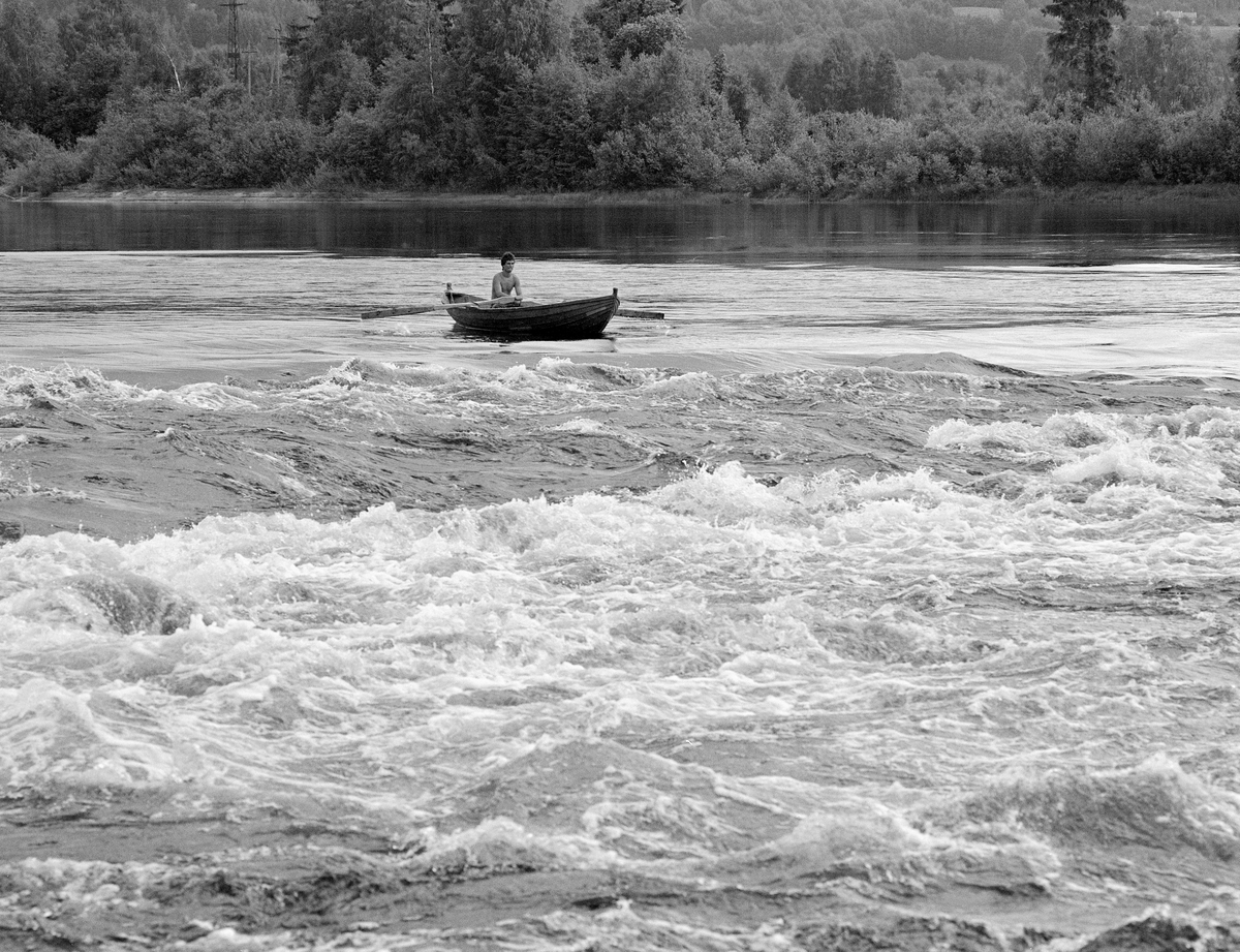 Fløterbåt ved Norsfossen i Glomma i begynnelsen av juli 1985.  Denne lokaliteten lå i den nordre delen av gamle Brandval herred, cirka 15 kilometer nord for Kongsvinger by.  De to fallene i Norsfossen har en høyde på drøyt tre meter ved lavvann.  Høydeforskjellen mellom ovenforliggende og nedenforliggende elveløp ble noe mindre i flomperioder.  Nedenfor Norsfossen fløt Glomma stille i cirka 8 kilometer, før vannmassene nådde neste fall, Gjølstadfossen.  Fotografiet viser en mann som var i ferd med å ro fra en stilleflytende del av elva, mot et mer opprørt stryk, der vannet gikk kvitskummende.  Mannen var en tømmerfløter i en av Glomma fellesfløtingsforenings robåter.  Han var en del av det laget som arbeidet med «sluttrensken».  Det innebar at de skulle få alt tømmer som hadde lagt seg mot elvebreddene eller strandet mot steinskjær eller på grunner ute i elveløpet til å flyte videre med strømmen.  Dette fløterlaget fulgte «bakenden» – de siste stokkene – nedover elveløpet.  Her har elva et par små fall etter å ha flytt stille og rolig i flere mil.  1985 var siste fløtingssesong i Glomma.  Da ble det bare fløtet fra Glomstadmora i Åmot kommune og sørover, og bare i hovedvassdraget.  Fløtingskvantumet var også svært beskjedent i forhold til det som hadde vært vanlig i Glomma noen tiår tidligere.  Følgelig ble det en kort sesong.  Utislaget startet så seint som 3. juni, og sluttrensken nådde Norsfossen etter en drøy måned, 4. juli, da dette bildet ble tatt.