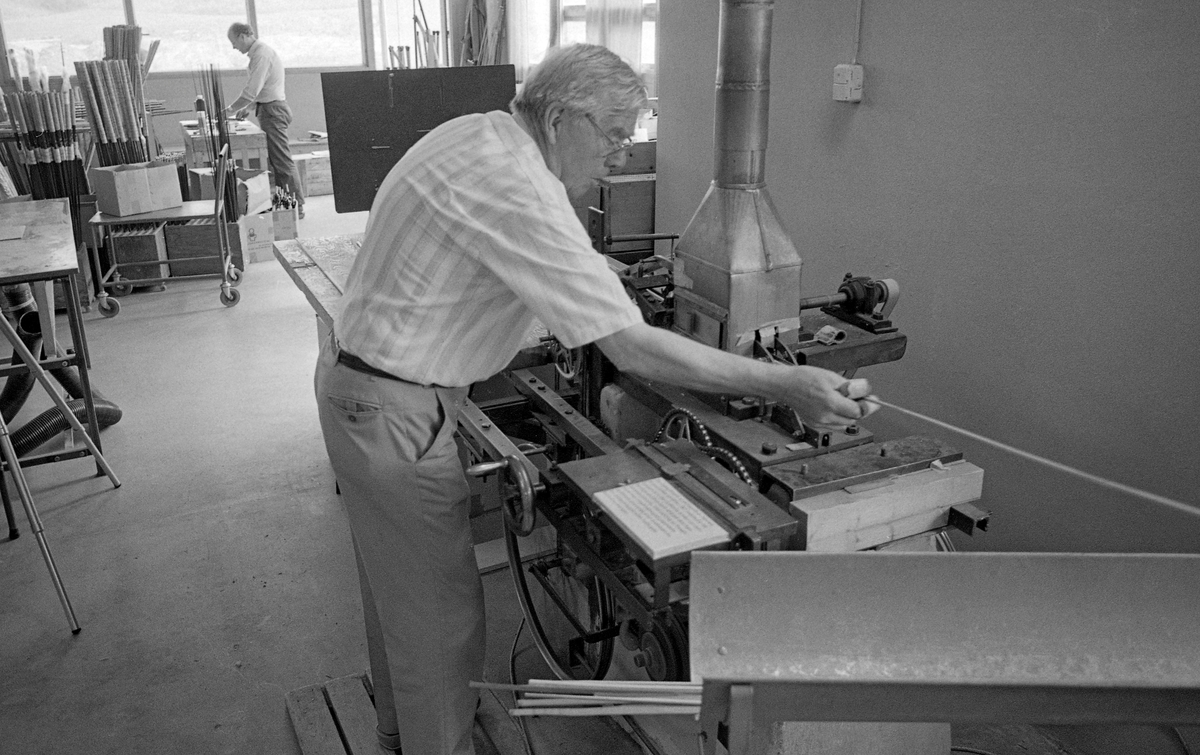 Industrigründeren Asbjørn Hørgård (1910-2001), fotografert i pruduksjonslokalene til bedriften han bygde opp i Trondheim.  Hørgård var særlig kjent for sine laminerte fiskestenger, såkalte splitcanestenger.  Veden i stengene var fra en bambusart som ble importert fra Østen.  Hørgård brukte også begrepet «tonkin», som antakelig henspilte på den sørøstasiatiske regionen råstoffet ble hentet fra.  Bambusen ble splittet i flere seksjoner (spildrer) og deretter limt sammen igjen.  Hørgård skrev følgende: «Den konstruksjonen som ble den seirende var den hvor spildrene ble høvlet i 60 graders vinkel og limt sammen til et enkeltbygget, sekskantet stangemne.  I grovere laksestenger ble det brukt dobbelt byggede stenger, dette på grunn av bambusspildrenes begrensede tykkelse.  Som konkurransestenger ble enhåndsstenger vesentlig hulbygget for å lette vekten og øke aksjonen».  Bambusstavene ble importert i 12 fots lengder og lagret i lang tid før bearbeidinga startet.  I fiskestangproduksjonen var det et poeng å pare stokkene slik at leddene, der de lange, seige fibrene startet og sluttet, ikke ble liggende mot hverandre.  Dette fotografiet viser Hørgård i aktivitet ved den maskinen han hadde konstruert for å kunne høvle bambusspildrene slik at de ble kone.  Bambusmaterialet hadde de mest spenstige fibrene i yteveden.  Derfor ble veden splitter og høvlet innenfra, først jevntjukke og deretter kone.  Hørgård forklarte dette slik:  «Høvlingene foregår på maskiner vi har laget selv.  Disse er meget nøyaktige og har uendelige muligheter for dimensjonering av spildrene og dermed også stangen.  Man kan starte åp en hvilken som helst dimensjon, kjøre en avsmalning på første fot, en annen avsmalning på den neste o.s.v.»  Fotografiet ble tatt i 1986.  Litt mer informasjon om Asbjørn Hørgård og den virksomheten han bygde opp som produsent og importør av utstyr for sportsfiske finnes under fanen «Opplysninger».