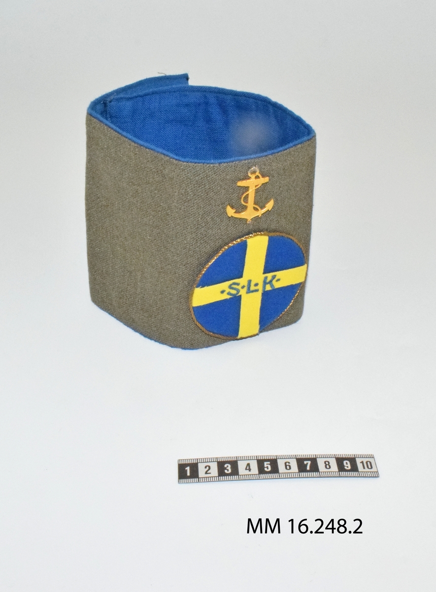 Armbindel för marinlotta. Rektangulärt tygstycke, dubbelt. Framstycket gråbrungrönt, bakstycket mellanblått. Bakstycket bildar blå passpoal runt det gråbrungröna stycket. I mitten på framstycket cirkelrunt emblem i tyg: Riksförbundet Sveriges lottakår: blå botten, gult kors med broderade bokstäver, blå: " . S . L . K .". Lagd gul metallpåsytt. Ovan emblemet gul metallankare fastsytt: flottans emblem.