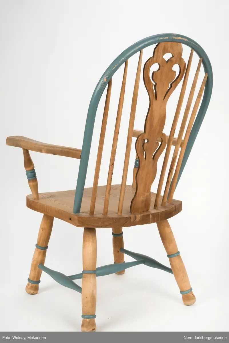 "Budal"inspirert stol, med ryggstø og armelner. Ryggstø er formet av en blåmalt buet list som holdes oppe av seks vertikale trespiler. Den midter, og syvende, spilen er en bred, utskjært list med gjennombrutt mønster, stiliserte hjertemønster. Armlenene er buet utover, og ben og detaljer har dreid dekor.