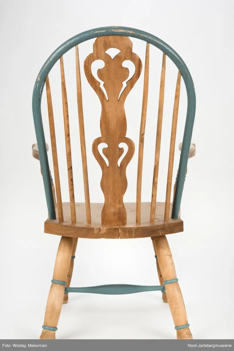 "Budal"inspirert stol, med ryggstø og armelner. Ryggstø er formet av en blåmalt buet list som holdes oppe av seks vertikale trespiler. Den midter, og syvende, spilen er en bred, utskjært list med gjennombrutt mønster, stiliserte hjertemønster. Armlenene er buet utover, og ben og detaljer har dreid dekor.