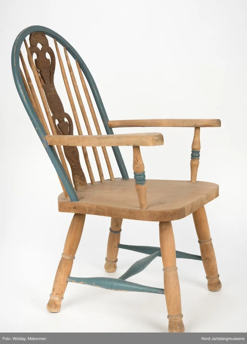 "Budal"inspirert stol, med ryggstø og armelner. Ryggstø er formet av en blåmalt buet list som holdes oppe av seks vertikale trespiler. Den midter, og syvende, spilen er en bred, utskjært list med gjennombrutt mønster, stiliserte hjertemønster. Armlenene er buet utover, og ben og detaljer har dreid dekor.