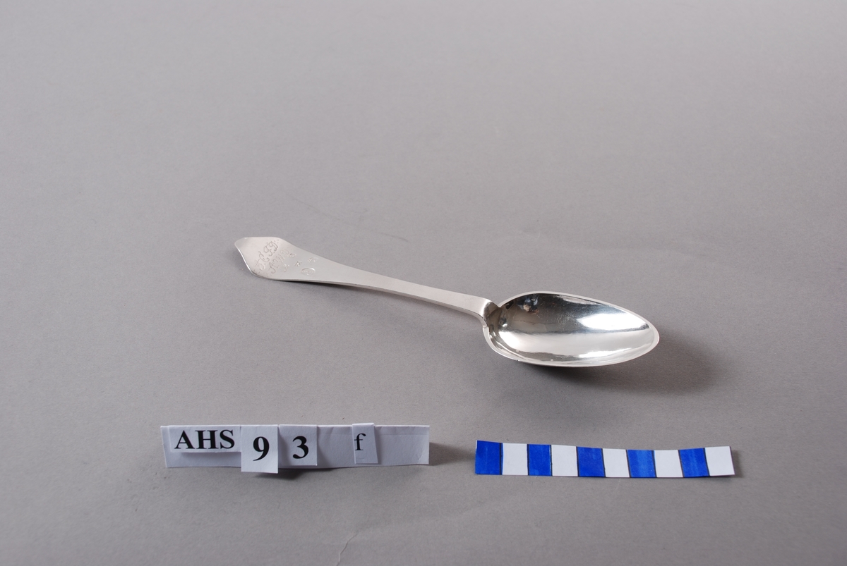 Spiseskje i sølv med stempel og inngraverte initialer. På baksiden er såvel blad som skaft meget rikt dekorert. Bladet har inngravert senrokokkobladverk, skaftet mer klassisistisk ornamentikk.