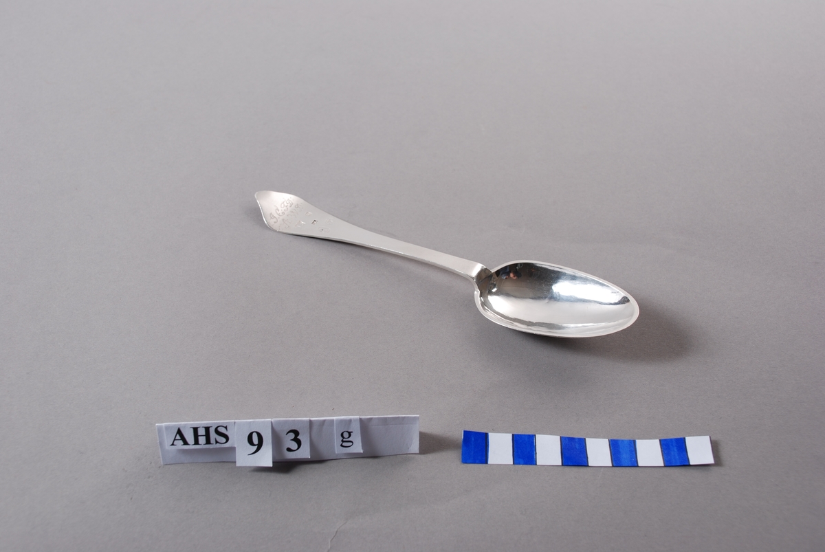 Spiseskje i sølv med stempel og inngraverte initialer. På baksiden er såvel blad som skaft meget rikt dekorert. Bladet har inngravert senrokokkobladverk, skaftet mer klassisistisk ornamentikk.