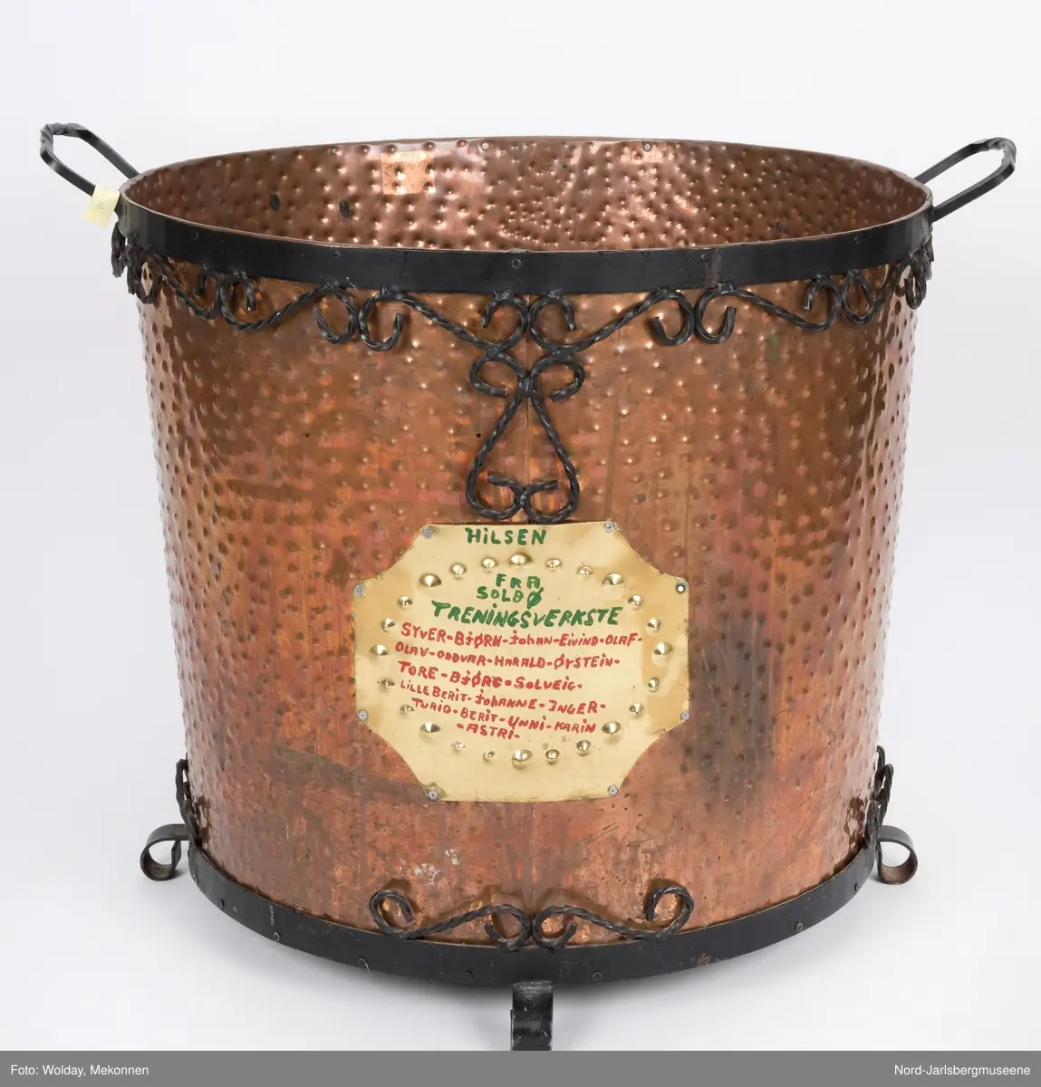 Stor kobberkjele i banket kobber med smijernsdekor i motstilte karnissformer. Smijernsbeslag rundt øvre kant og rundt bunnen, fire små smijernsben. På fronten er festet en messingplate med rød og grønn påskrift