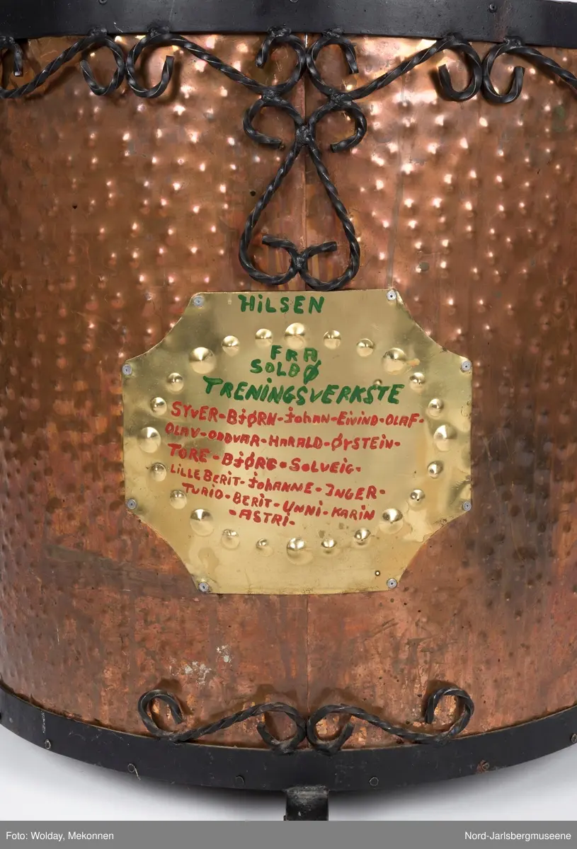 Stor kobberkjele i banket kobber med smijernsdekor i motstilte karnissformer. Smijernsbeslag rundt øvre kant og rundt bunnen, fire små smijernsben. På fronten er festet en messingplate med rød og grønn påskrift