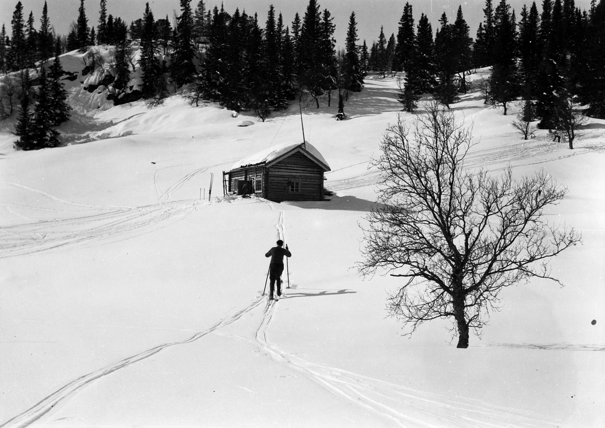 Mann gående på ski mot fjellhytte på Heddevasstulen. To par ski stående på utsiden av hytta. Skispor i lia ved siden av hytta. Hytta er oppført i laft.